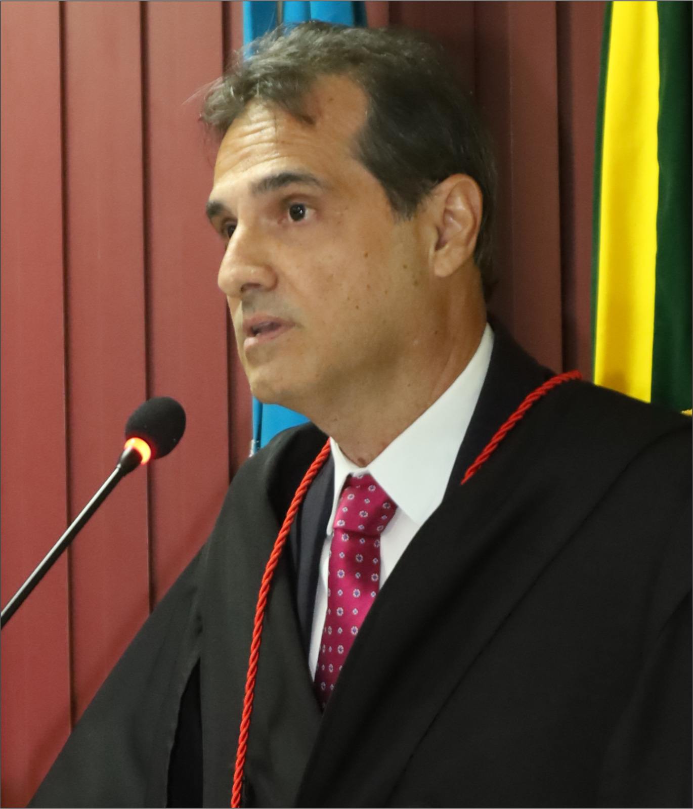 Fotografia de um homem usando veste talar preta com detalhes vermelhos, camisa branca e gravata vermelha, posicionado ao lado de um microfone durante discurso. Fotografia de um homem usando veste talar preta com detalhes vermelhos, camisa branca e gravata vermelha, posicionado ao lado de um microfone durante discurso.