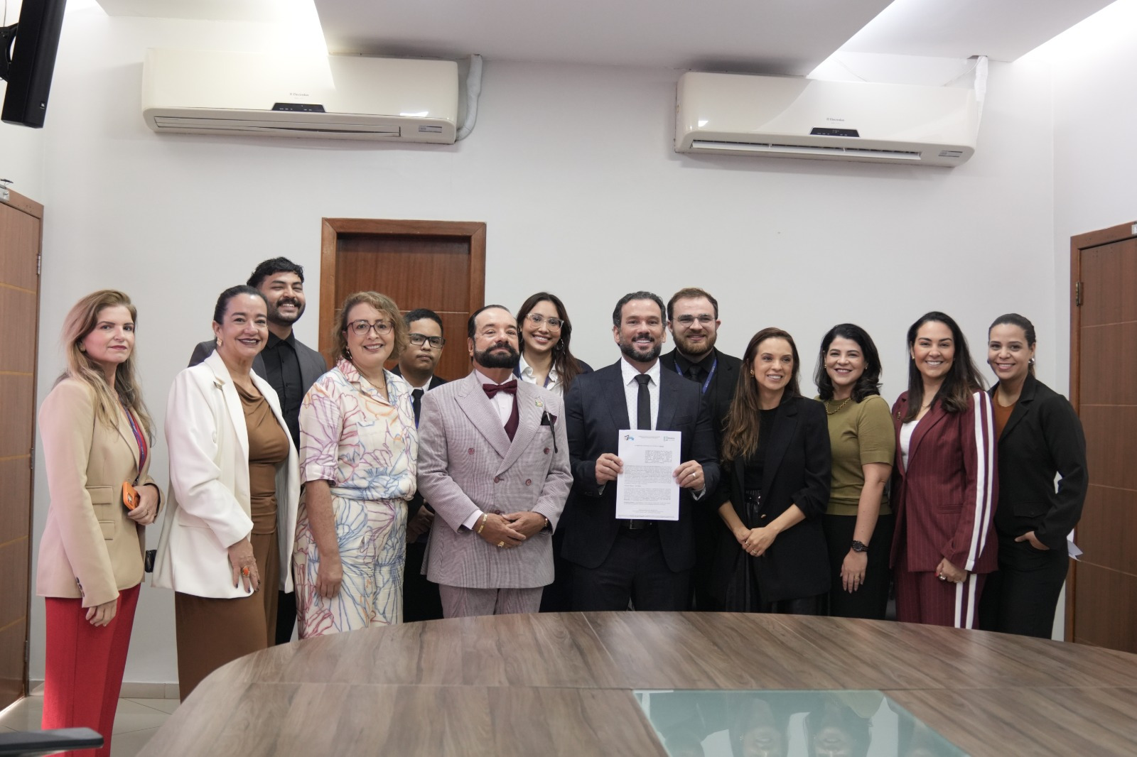 Imagem colorida mostra um grupo de pessoas posando para foto em uma sala de reunião. Ao centro, um homem de terno escuro segura um documento. Ao lado dele estão representantes das instituições envolvidas, todos sorrindo para a câmera. No ambiente, há uma mesa ampla em primeiro plano, portas de madeira e aparelhos de ar-condicionado na parede.