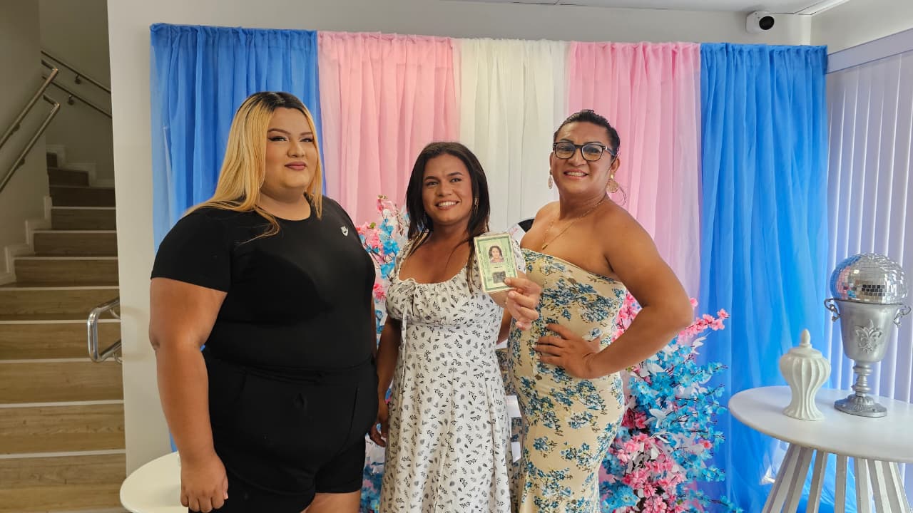 Três pessoas posam sorrindo em frente ao painel decorado do evento. Uma delas segura um documento de identificação.