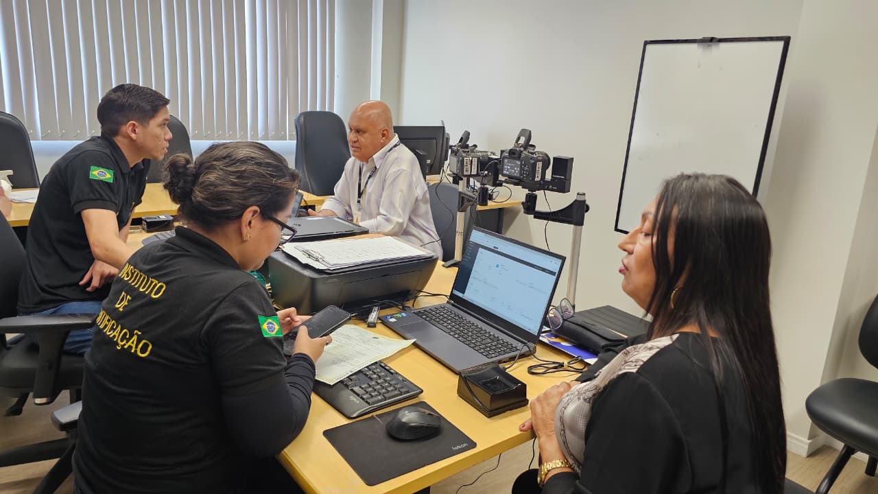 Equipe de atendimento atua em uma sala com computadores, impressora e equipamentos. Pessoas conversam e conferem informações durante os serviços do mutirão.