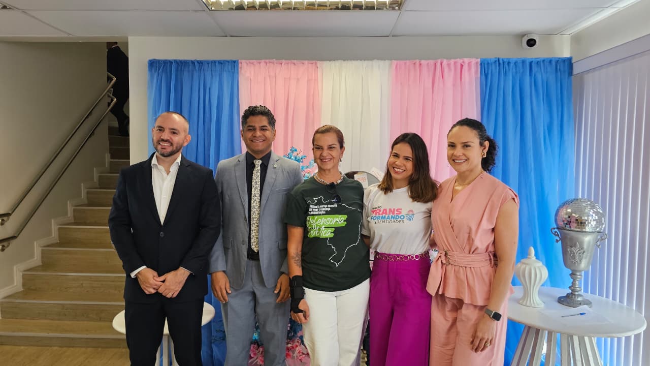 Cinco pessoas posam para foto em frente a um painel decorado nas cores azul, rosa e branco. Uma delas usa camiseta com a identificação do Mutirão TransFormando Identidades.
