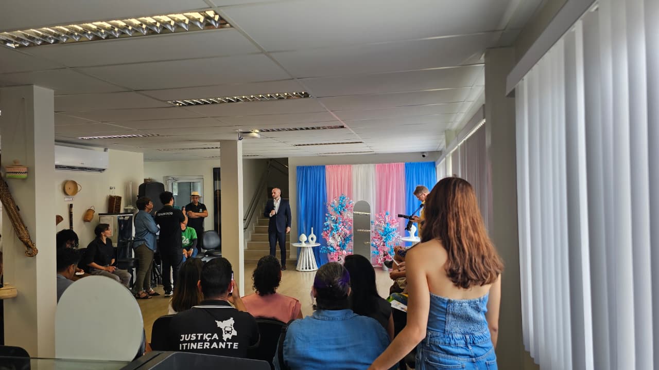 Público acompanha uma fala durante o Mutirão TransFormando Identidades. Ao fundo, há decoração nas cores azul, rosa e branco, além de pessoas da equipe da Justiça Itinerante.
