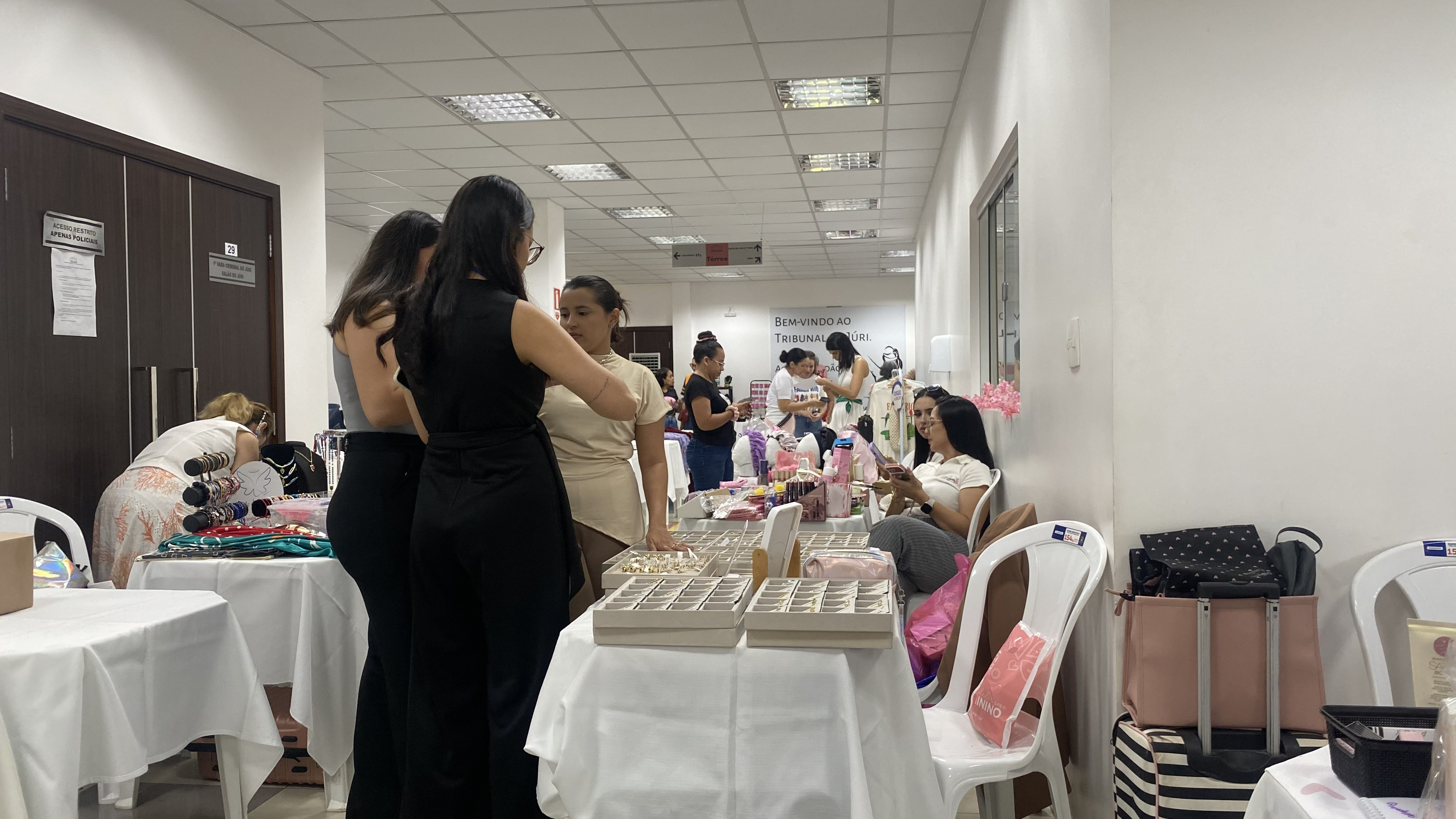 Vista lateral de um corredor com mesas brancas alinhadas expondo joias e acessórios em bandejas organizadoras. Várias mulheres conversam e olham os produtos expostos. Vista lateral de um corredor com mesas brancas alinhadas expondo joias e acessórios em bandejas organizadoras. Várias mulheres conversam e olham os produtos expostos.
