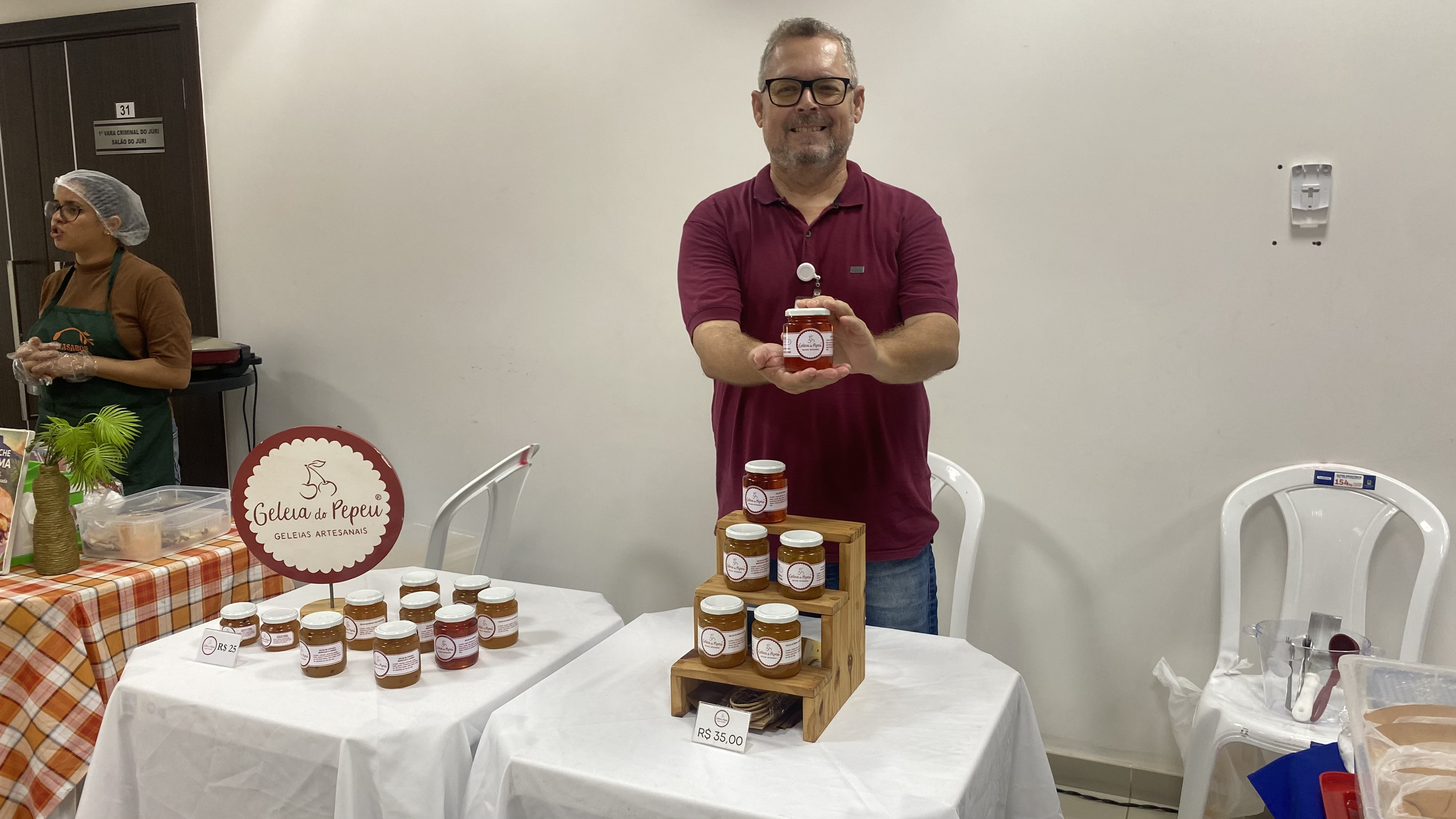 Um homem sorridente de óculos e camisa polo vinho posa segurando um pequeno pote de geleia. À sua frente, sobre mesas brancas, há vários outros potes organizados em um pequeno suporte de madeira e um painel redondo com a marca do produto. Um homem sorridente de óculos e camisa polo vinho posa segurando um pequeno pote de geleia. À sua frente, sobre mesas brancas, há vários outros potes organizados em um pequeno suporte de madeira e um painel redondo com a marca do produto.