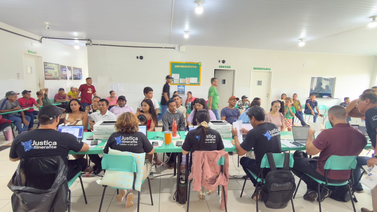 Foto colorida mostra os atendimentos da Justiça Itinerante em um espaço amplo e iluminado. Servidores realizam atendimentos em mesas com computadores e impressoras, enquanto moradores aguardam atendimento sentados ou em fila Foto colorida mostra os atendimentos da Justiça Itinerante em um espaço amplo e iluminado. Servidores realizam atendimentos em mesas com computadores e impressoras, enquanto moradores aguardam atendimento sentados ou em fila