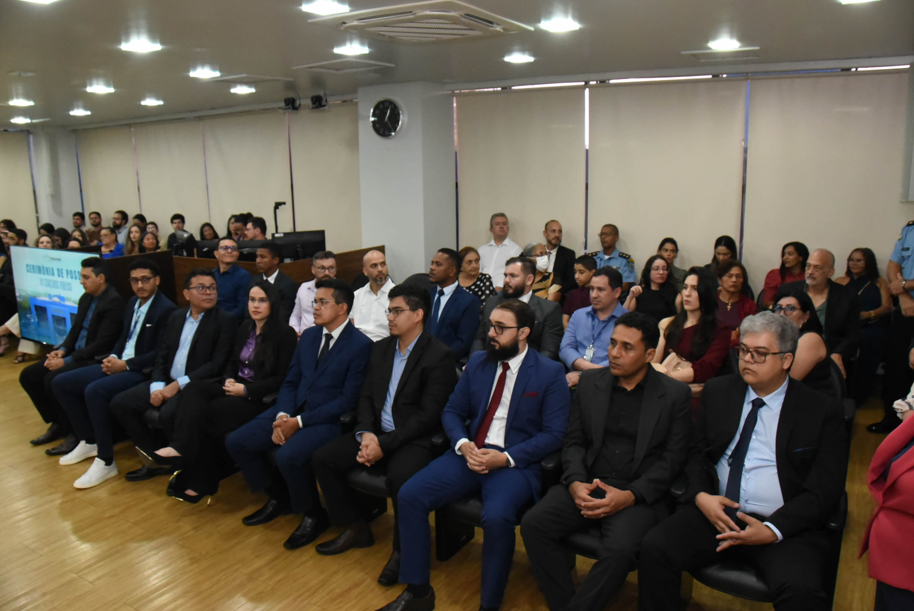 Grupo de pessoas em uma cerimônia de posse, sentadas em cadeiras, com expressões formais e atenção voltada para o apresentador.