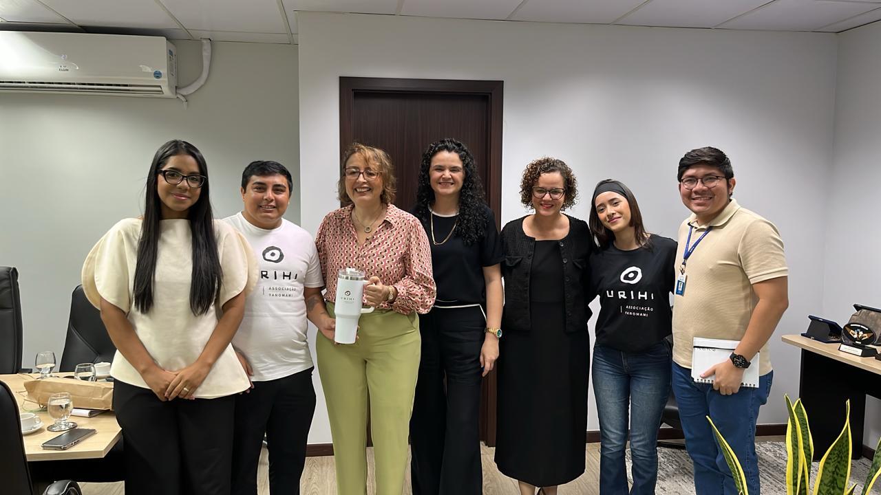 Sete pessoas estão sorrindo lado a lado em uma sala de reuniões comercial, com duas delas vestindo camisetas pretas e brancas da Associação Yanomami Urihi. Sete pessoas estão sorrindo lado a lado em uma sala de reuniões comercial, com duas delas vestindo camisetas pretas e brancas da Associação Yanomami Urihi.