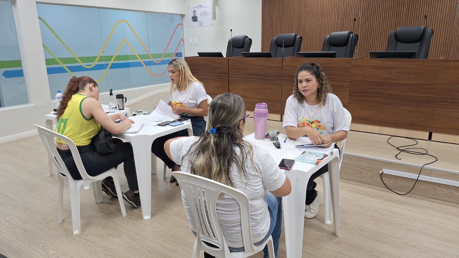 Foto 2: Imagem colorida mostra mulheres reunidas em duas mesas brancas de plástico em um auditório judiciário, conversando e preenchendo documentos de atendimento.