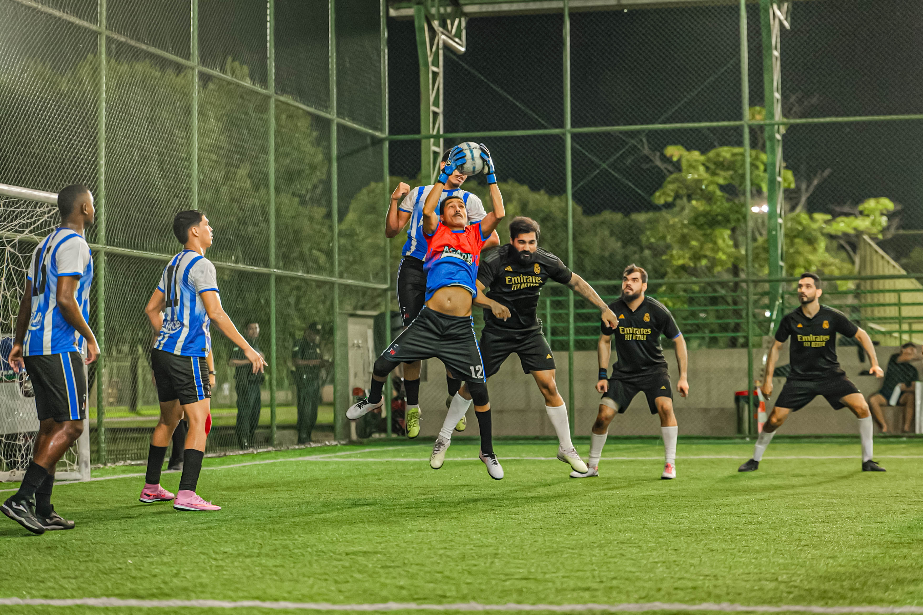 Foto 2: Imagem colorida mostra uma partida de futebol society à noite, onde um goleiro de uniforme vermelho e azul pula alto para segurar a bola no ar em meio a jogadores rivais de preto. Foto 2: Imagem colorida mostra uma partida de futebol society à noite, onde um goleiro de uniforme vermelho e azul pula alto para segurar a bola no ar em meio a jogadores rivais de preto.