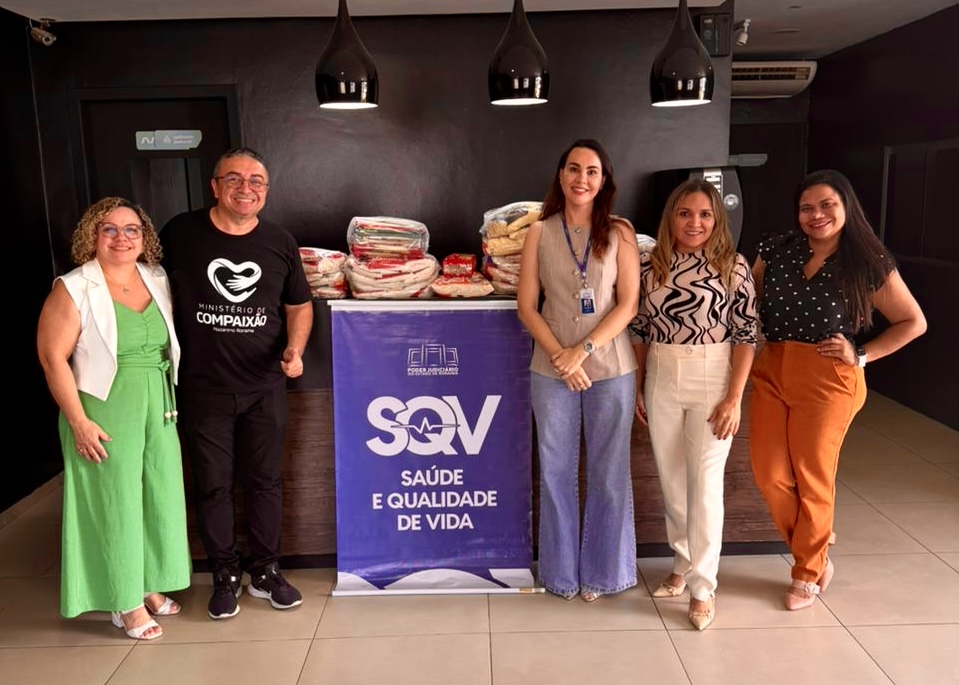 Foto 3: Imagem colorida mostra cinco pessoas posando para foto ao lado de um banner roxo com a inscrição "SQV - Saúde e Qualidade de Vida", tendo ao fundo e mantimentos sobre um balcão. Foto 3: Imagem colorida mostra cinco pessoas posando para foto ao lado de um banner roxo com a inscrição "SQV - Saúde e Qualidade de Vida", tendo ao fundo e mantimentos sobre um balcão.