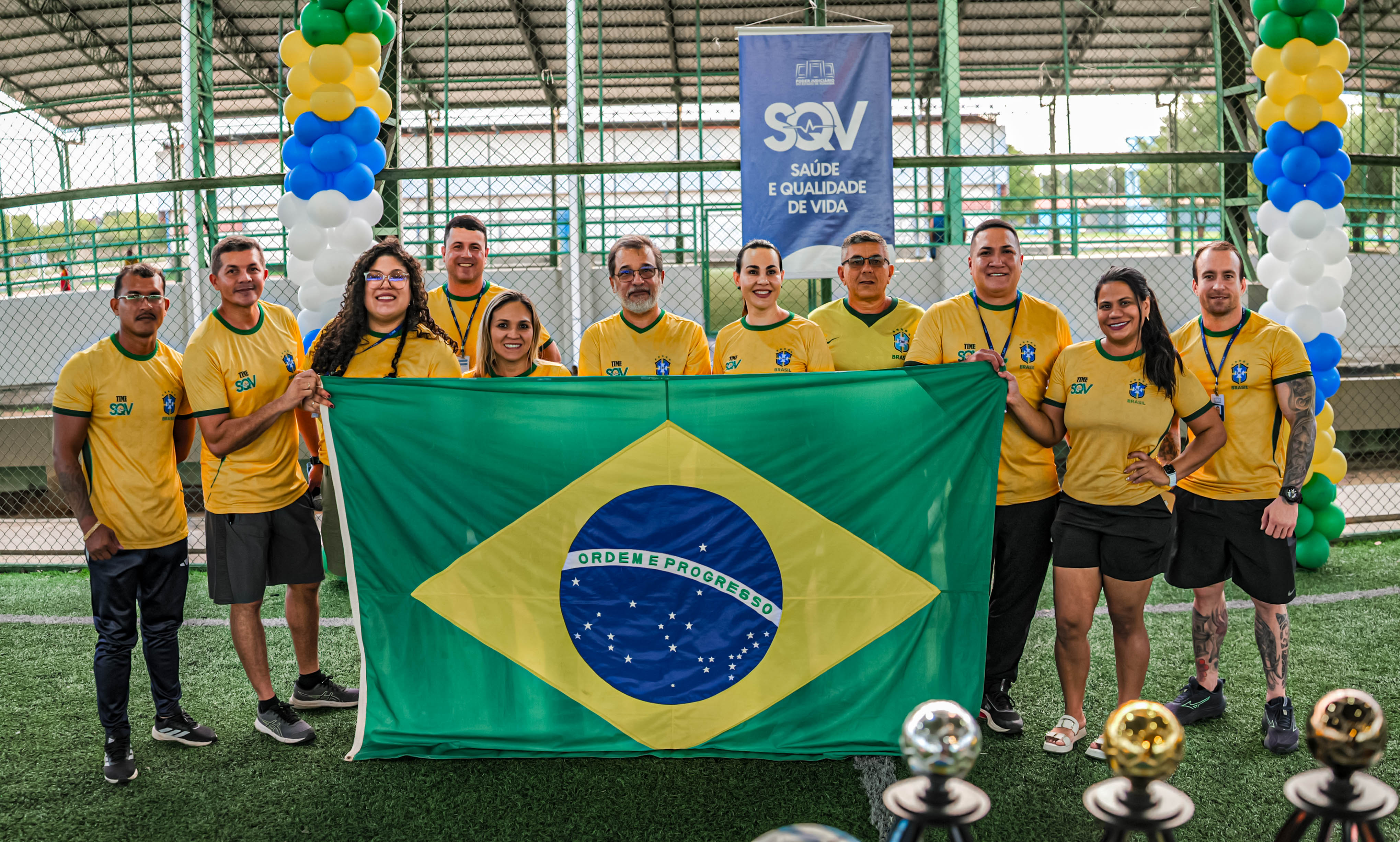 Foto 4: Imagem colorida mostra um grupo de pessoas vestindo camisas amarelas da seleção brasileira posando em uma quadra esportiva enquanto segura uma grande bandeira do Brasil esticada à frente. Foto 4: Imagem colorida mostra um grupo de pessoas vestindo camisas amarelas da seleção brasileira posando em uma quadra esportiva enquanto segura uma grande bandeira do Brasil esticada à frente.