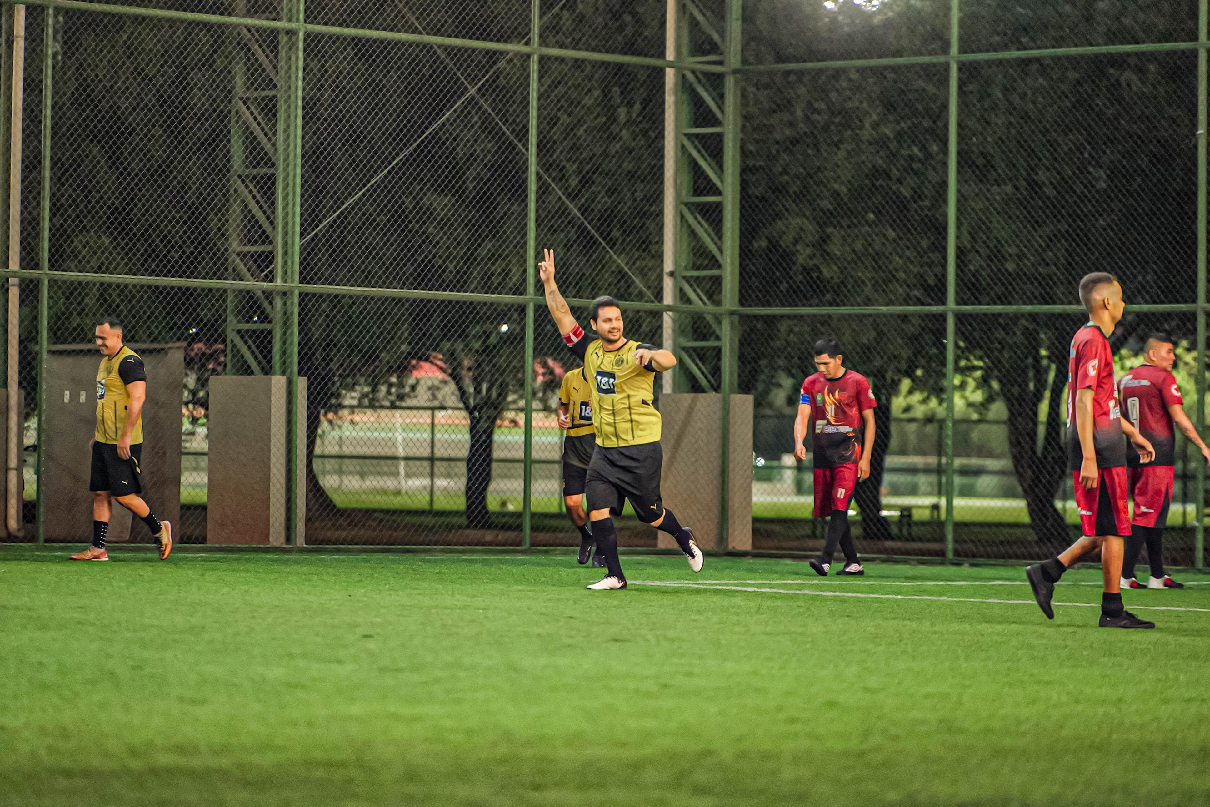 Foto 5: Imagem colorida mostra uma partida de futebol society à noite; ao centro, um jogador de uniforme amarelo e preto comemora correndo com o braço levantado fazendo um sinal de "2" com os dedos. Foto 5: Imagem colorida mostra uma partida de futebol society à noite; ao centro, um jogador de uniforme amarelo e preto comemora correndo com o braço levantado fazendo um sinal de "2" com os dedos.