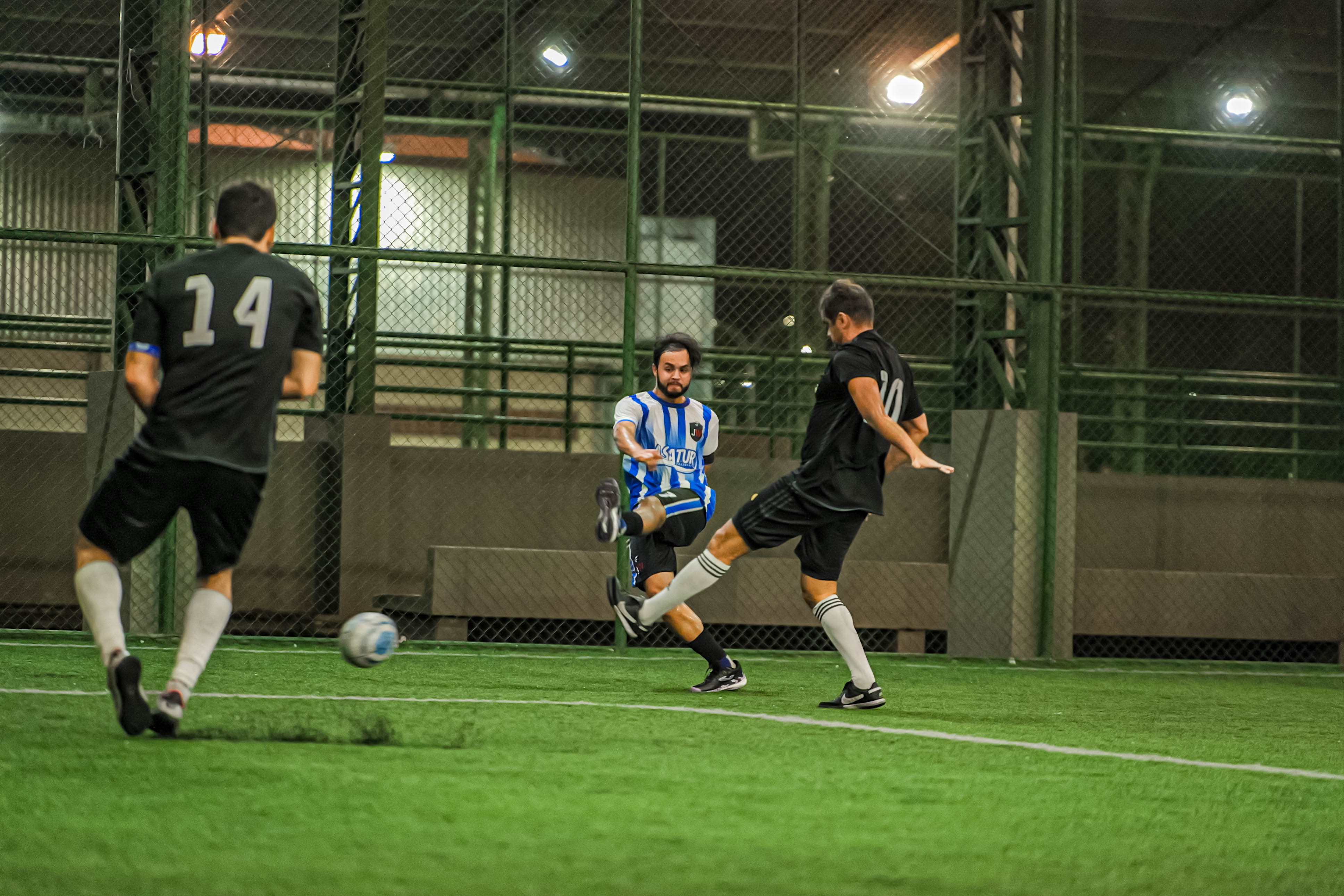 Foto 6: Imagem colorida mostra uma partida de futebol society à noite; um jogador de uniforme listrado em azul e branco chuta a bola enquanto é marcado por adversários de uniforme preto. Foto 6: Imagem colorida mostra uma partida de futebol society à noite; um jogador de uniforme listrado em azul e branco chuta a bola enquanto é marcado por adversários de uniforme preto.
