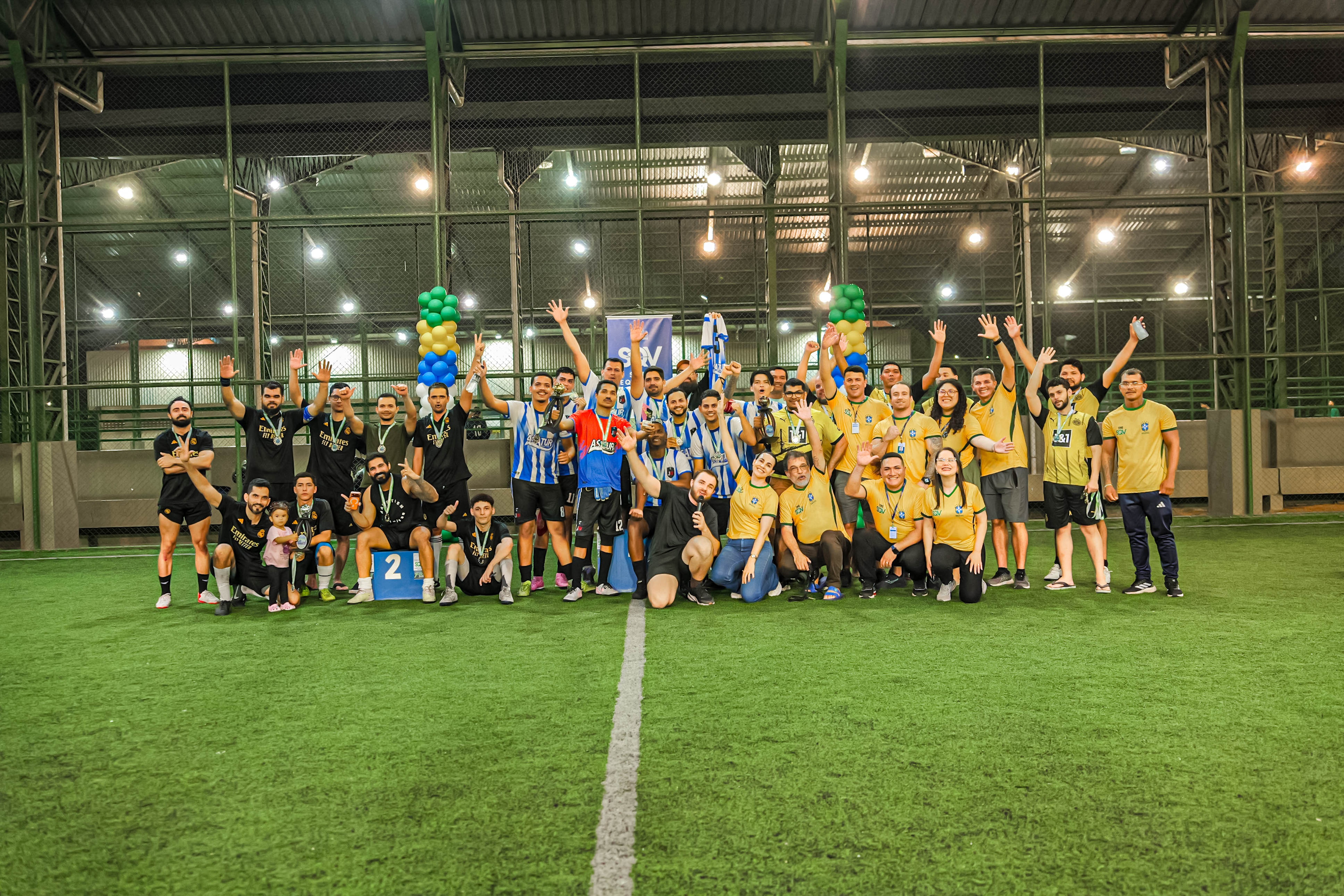 Foto 7: Imagem colorida mostra um grande grupo de pessoas e jogadores reunidos no centro de uma quadra de futebol society acenando e comemorando o fim de um torneio esportivo noturno. Foto 7: Imagem colorida mostra um grande grupo de pessoas e jogadores reunidos no centro de uma quadra de futebol society acenando e comemorando o fim de um torneio esportivo noturno.