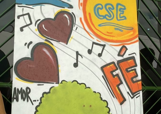Foto 2: Imagem colorida mostra uma pintura em grafite contendo dois corações marrons, notas musicais, um sol amarelo com a sigla "CSE", uma árvore verde e as palavras "FÉ" e "AMOR”. Foto 2: Imagem colorida mostra uma pintura em grafite contendo dois corações marrons, notas musicais, um sol amarelo com a sigla "CSE", uma árvore verde e as palavras "FÉ" e "AMOR”.