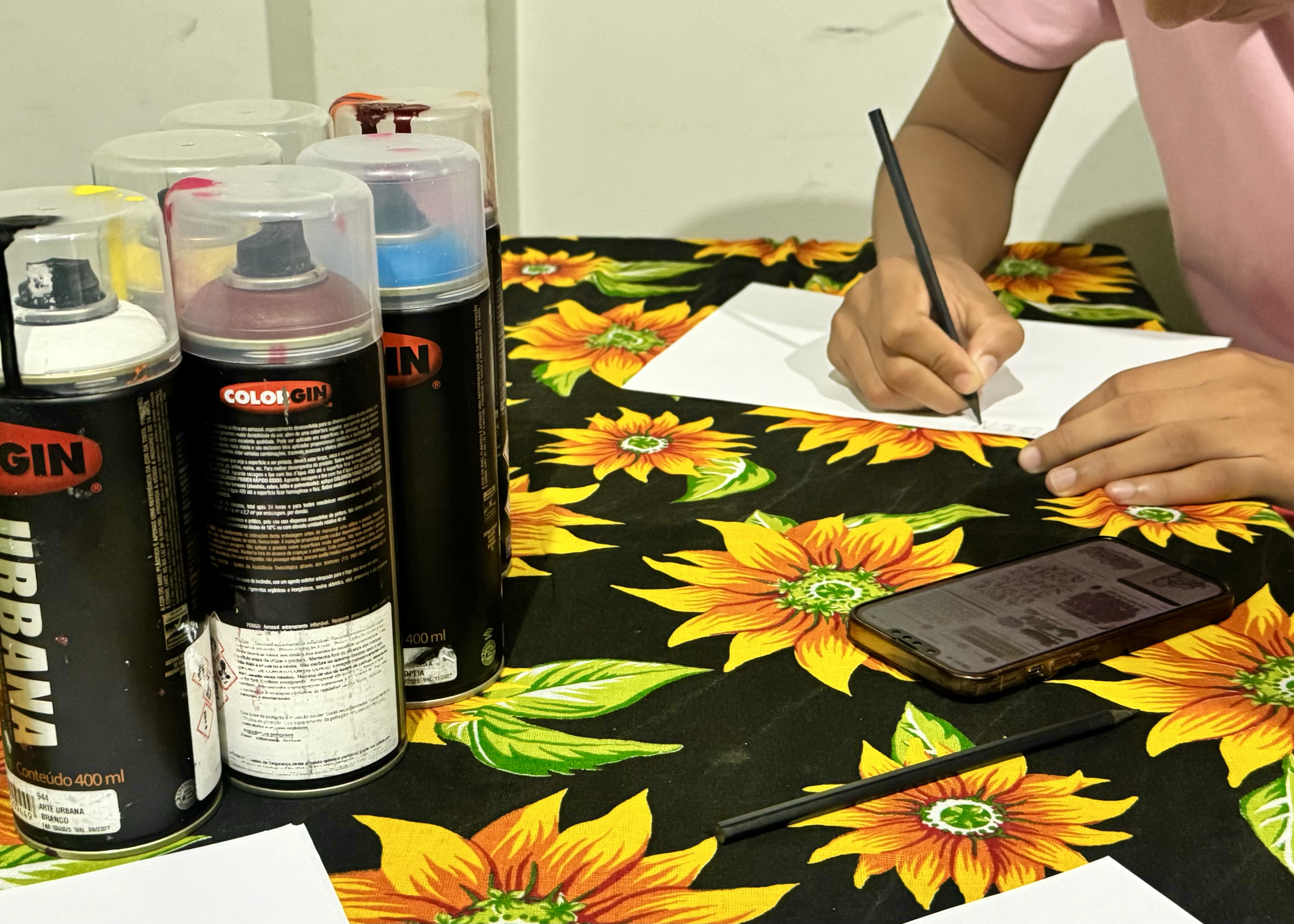 Foto 3: Imagem colorida mostra uma pessoa desenhando com lápis em uma folha de papel sobre uma mesa com toalha de girassóis, cercada por várias latas de tinta spray e um celular. Foto 3: Imagem colorida mostra uma pessoa desenhando com lápis em uma folha de papel sobre uma mesa com toalha de girassóis, cercada por várias latas de tinta spray e um celular.