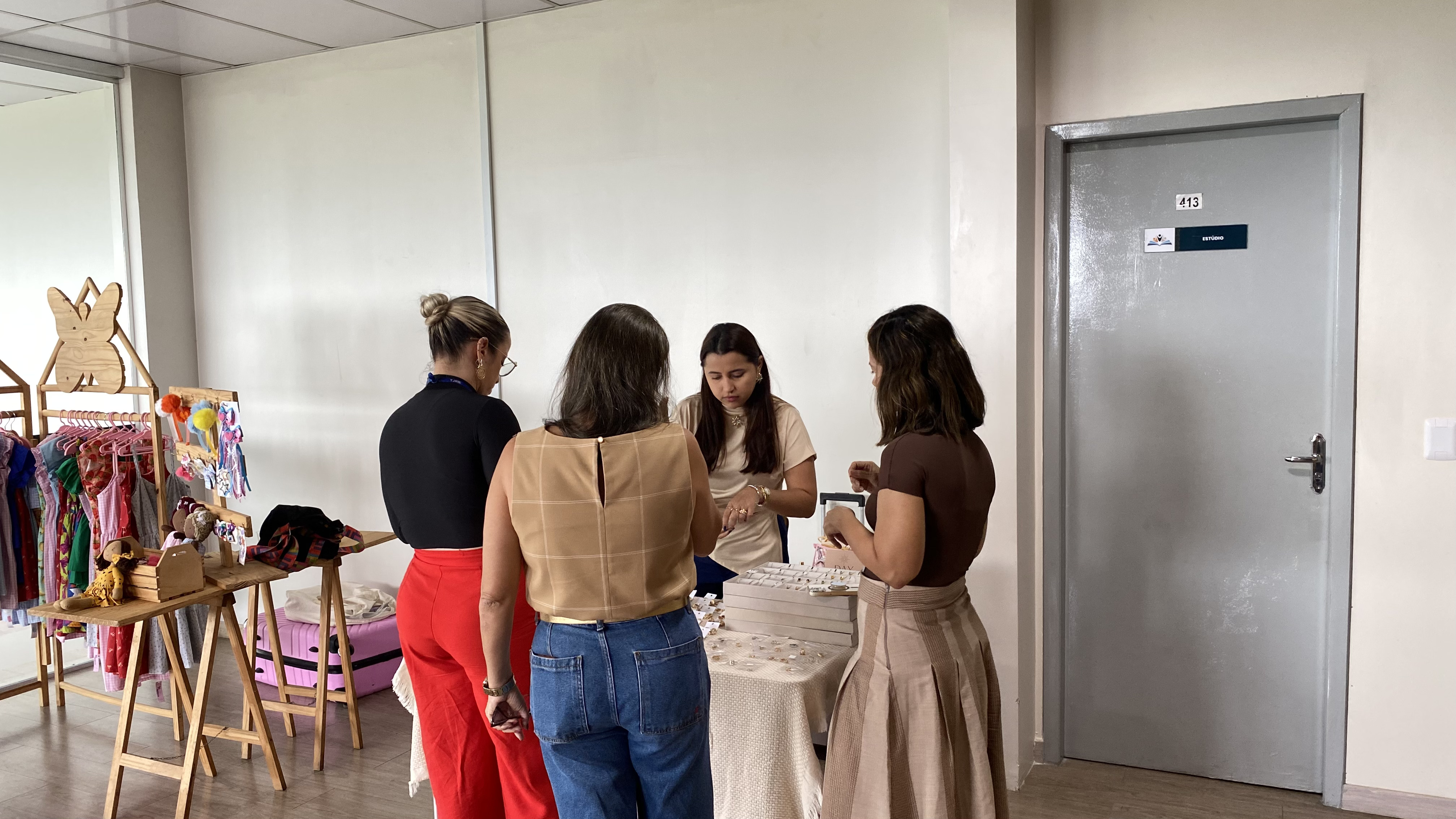 Fotografia de um estande na feira de empreendedorismo. No centro, uma expositora atende três mulheres ao redor de uma mesa com bandejas de acessórios. À esquerda, há suportes de madeira com roupas infantis e laços expostos. À direita, vê-se uma porta cinza fechada. Fotografia de um estande na feira de empreendedorismo. No centro, uma expositora atende três mulheres ao redor de uma mesa com bandejas de acessórios. À esquerda, há suportes de madeira com roupas infantis e laços expostos. À direita, vê-se uma porta cinza fechada.