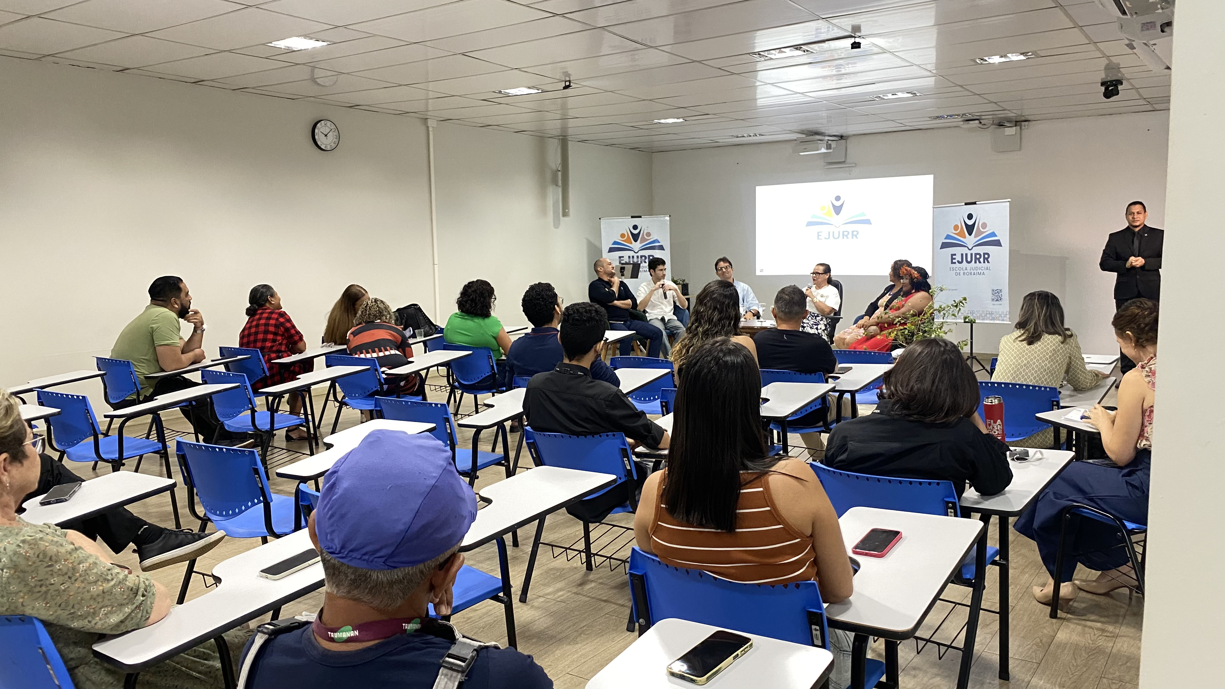 Vista geral de um auditório onde o público, sentado em cadeiras azuis com prancheta, assiste à roda de conversa. Ao fundo da sala, os participantes estão reunidos no palco em frente a banners e uma tela de projeção com a marca da EJURR. No canto direito, um intérprete de Libras acompanha o evento de pé. Vista geral de um auditório onde o público, sentado em cadeiras azuis com prancheta, assiste à roda de conversa. Ao fundo da sala, os participantes estão reunidos no palco em frente a banners e uma tela de projeção com a marca da EJURR. No canto direito, um intérprete de Libras acompanha o evento de pé.