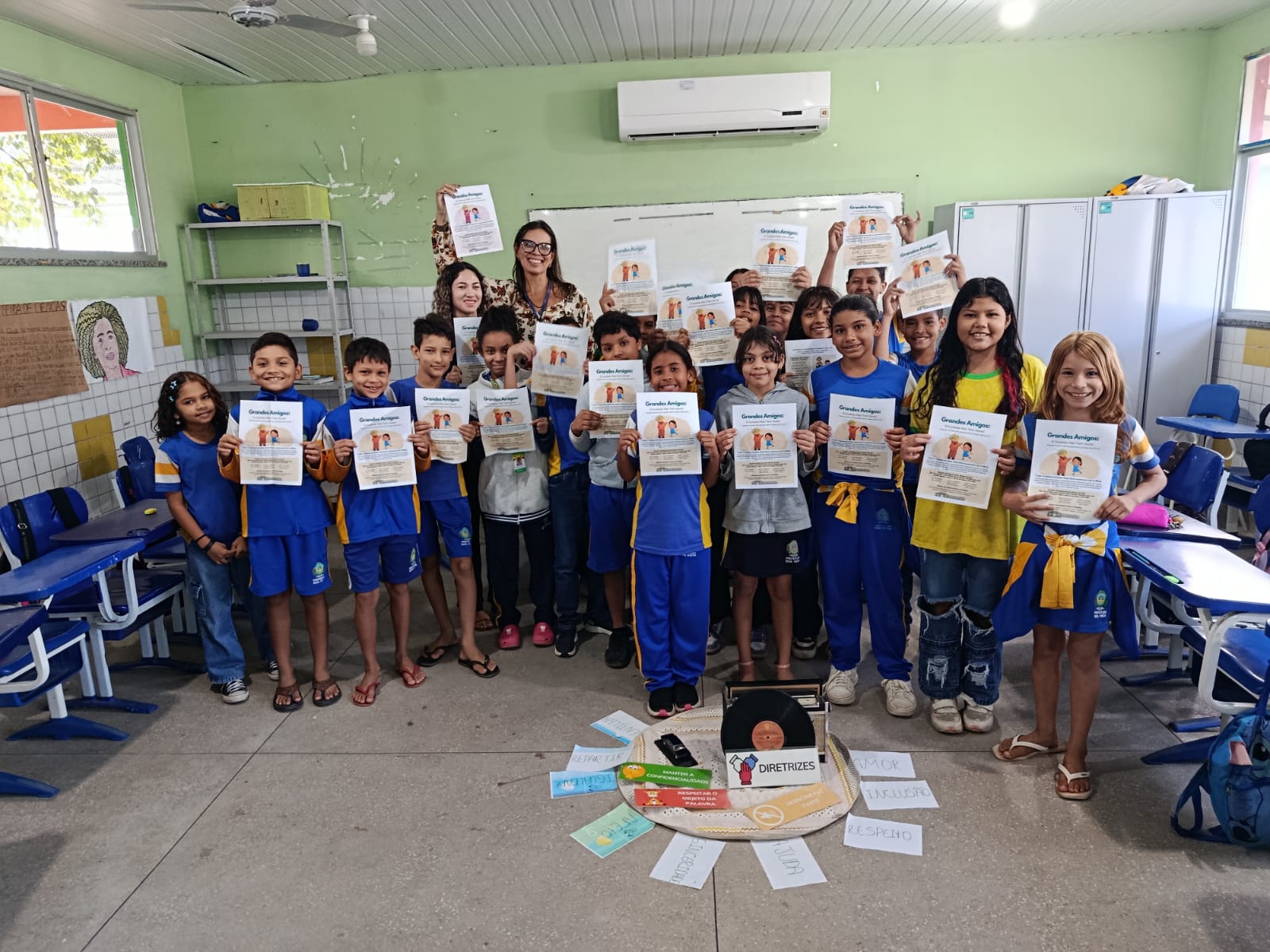 Fotografia de um grupo de estudantes do ensino fundamental e educadoras reunidos em uma sala de aula. Eles sorriem e mostram o livreto informativo "Grandes Amigos". No chão, à frente do grupo, há um arranjo com cartões coloridos contendo palavras como "Respeito", "Amor" e "Inclusão". Fotografia de um grupo de estudantes do ensino fundamental e educadoras reunidos em uma sala de aula. Eles sorriem e mostram o livreto informativo "Grandes Amigos". No chão, à frente do grupo, há um arranjo com cartões coloridos contendo palavras como "Respeito", "Amor" e "Inclusão".