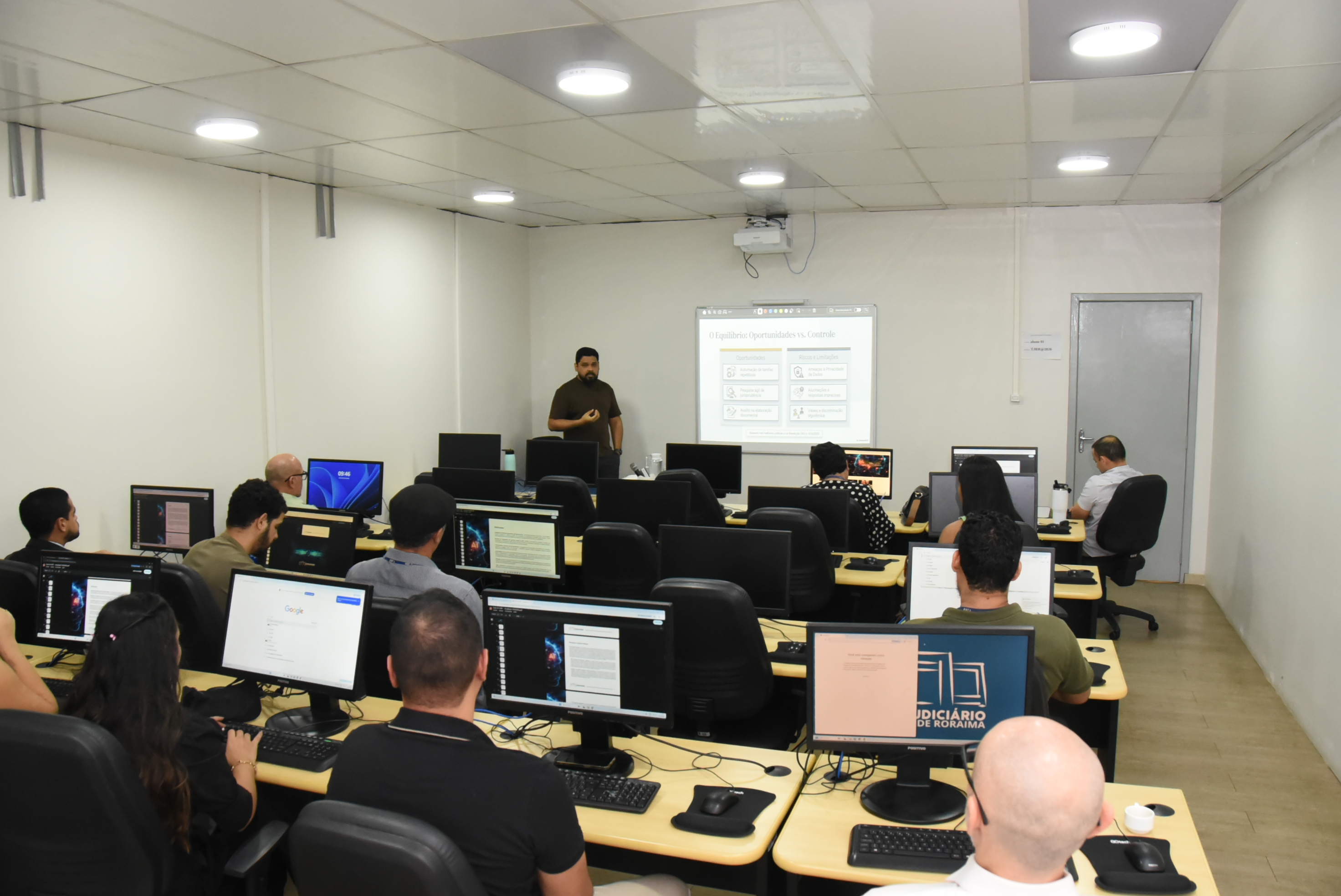 Foto 2: Imagem colorida mostra uma sala de informática onde várias pessoas estão sentadas em frente a computadores assistindo a uma apresentação de um instrutor de pé ao fundo, ao lado de uma tela de projeção. Foto 2: Imagem colorida mostra uma sala de informática onde várias pessoas estão sentadas em frente a computadores assistindo a uma apresentação de um instrutor de pé ao fundo, ao lado de uma tela de projeção.