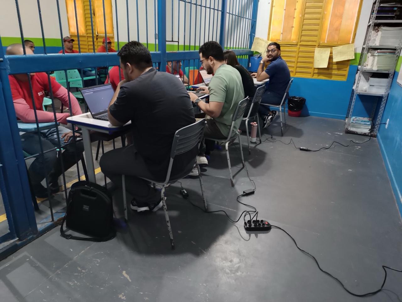 Imagem colorida mostra equipe técnica em atendimento com uso de notebooks, ao lado de grades azuis, enquanto pessoas privadas de liberdade aguardam sentadas do outro lado.