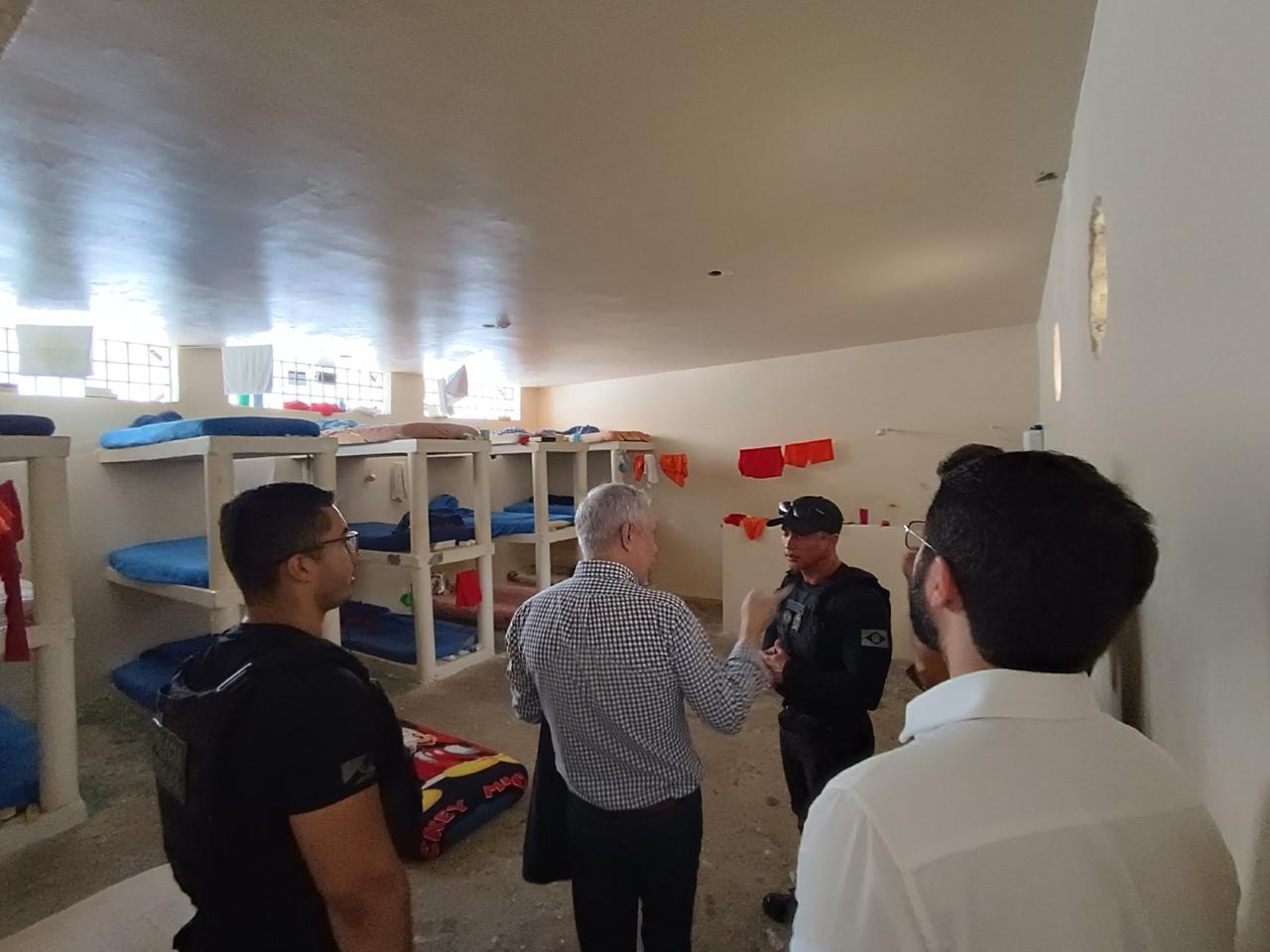 Imagem colorida mostra representantes do Judiciário e agentes de segurança conversando dentro de um alojamento com beliches e colchões azuis em uma unidade prisional.