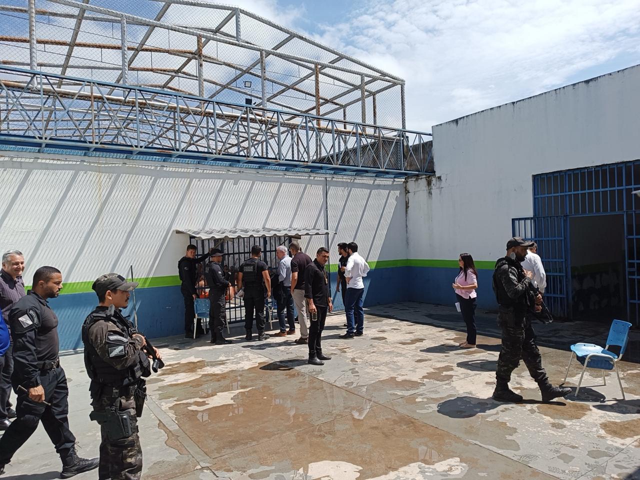 Imagem colorida mostra pátio externo de unidade prisional, com policiais penais armados e representantes institucionais reunidos durante visita técnica de fiscalização.