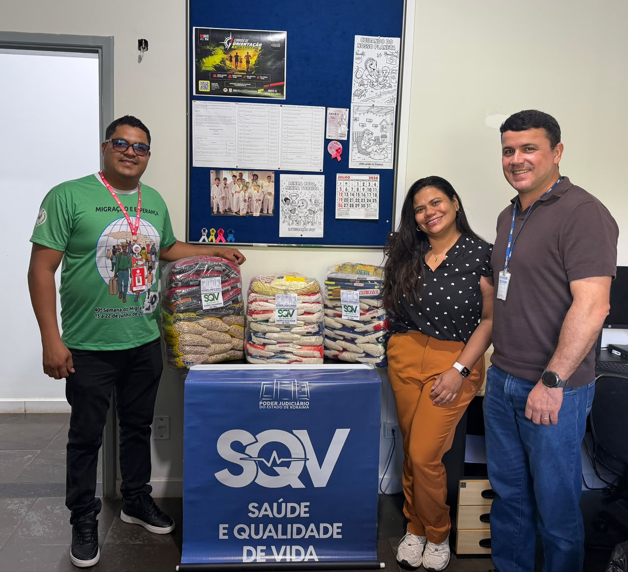 Três pessoas sorrindo de pé em um escritório ao lado de várias pilhas de alimentos não perecíveis arrecadados, posicionados atrás de uma placa azul da SQV (Saúde e Qualidade de Vida).