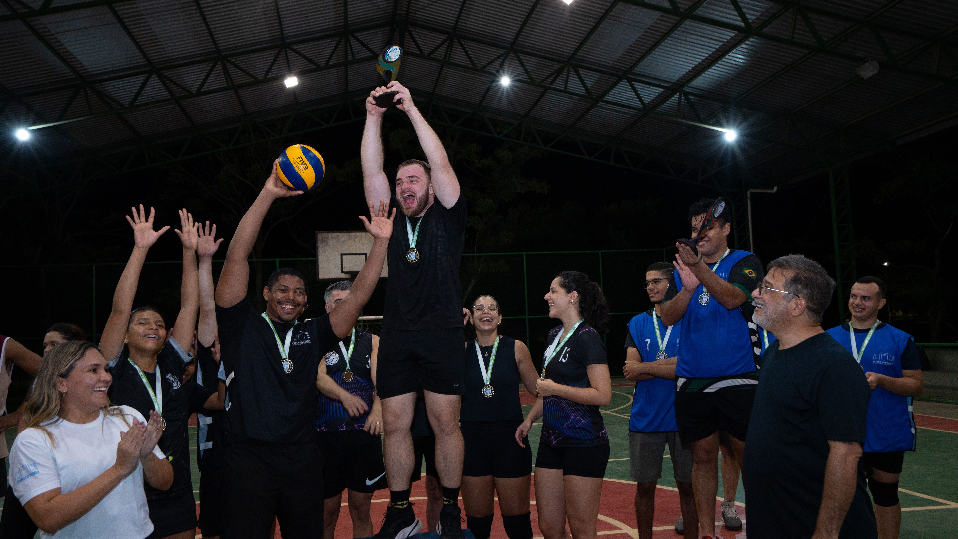 Jogador do Time Preto erguido no pódio, sorrindo e levantando o troféu de campeão, enquanto seus companheiros de equipe comemoram ao redor na quadra.