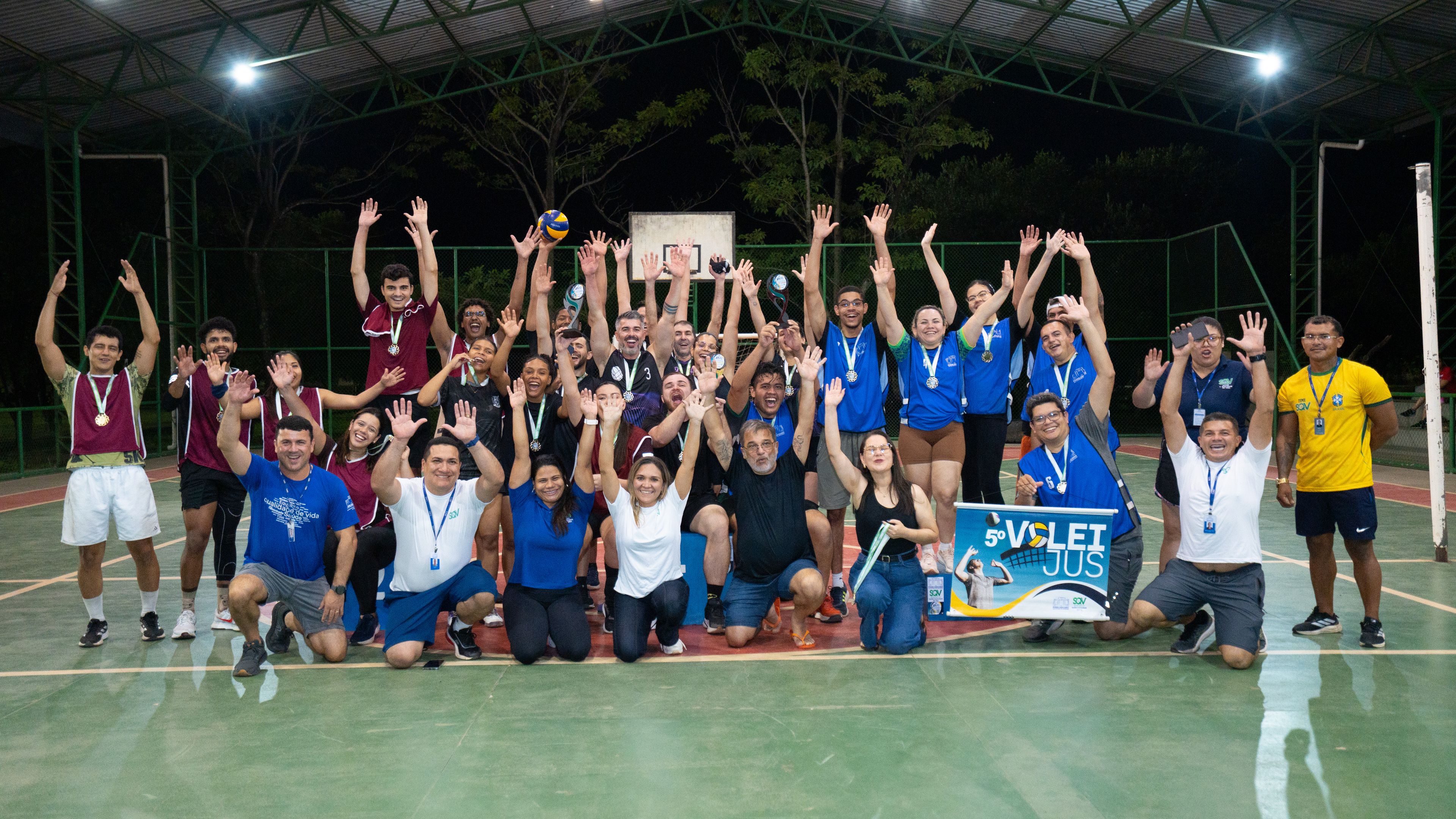 Foto oficial de todos os participantes do torneio reunidos na quadra, sorrindo e comemorando com os braços levantados. No canto inferior direito, uma pessoa segura o banner do "5º VôleiJus".
