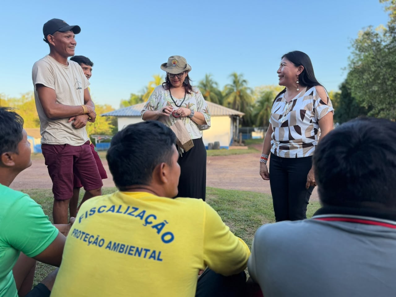 Foto 2: Imagem colorida mostra um grupo de pessoas conversando em uma área aberta gramada ao final da tarde. No primeiro plano, de costas, um homem usa camiseta amarela com a inscrição "FISCALIZAÇÃO PROTEÇÃO AMBIENTAL". Ao fundo, duas mulheres e outros homens interagem sorrindo, com uma casa simples e árvores sob o céu azul.