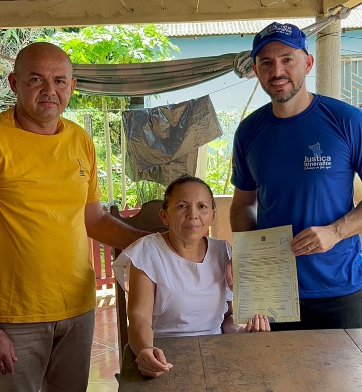 Mulher sentada exibe certidão ao lado de familiar e o juiz subistituto da Justiça Itinerante, após atendimento domiciliar. Mulher sentada exibe certidão ao lado de familiar e o juiz subistituto da Justiça Itinerante, após atendimento domiciliar.