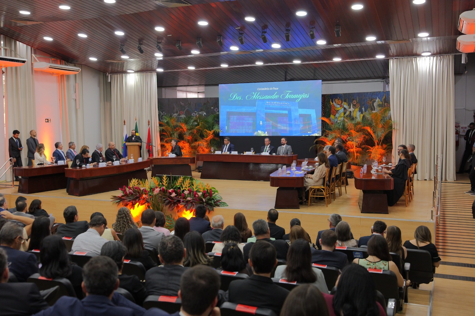 Imagem colorida mostra visão ampla do auditório durante a sessão solene de posse, com autoridades compondo a mesa, público presente e painel ao fundo com a identificação da cerimônia.