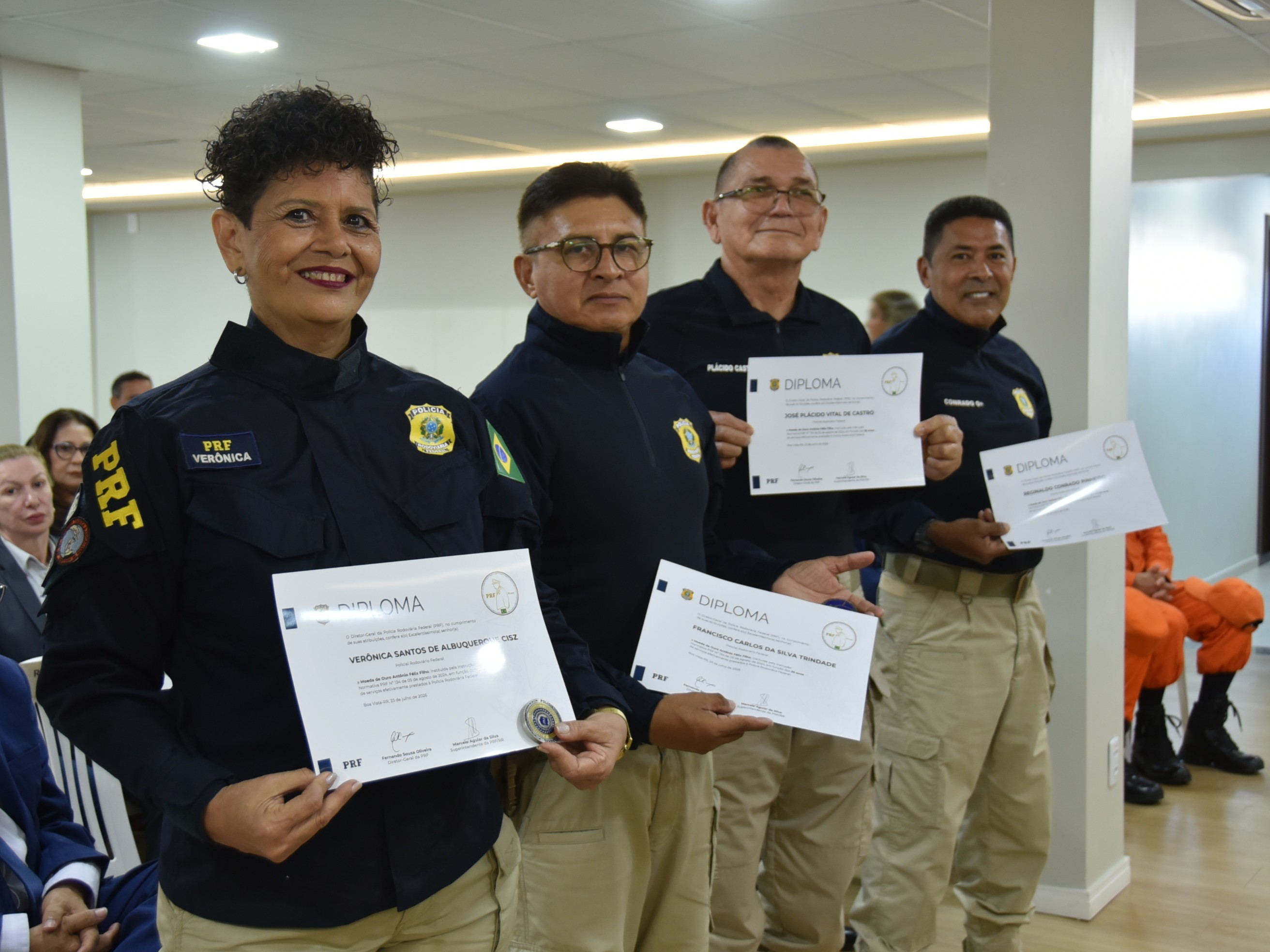 Imagem colorida mostra quatro policiais rodoviários federais veteranos posando com diplomas e medalhas recebidos durante a cerimônia de homenagem. À frente está a inspetora Verônica Cisz, acompanhada por outros três integrantes da instituição.