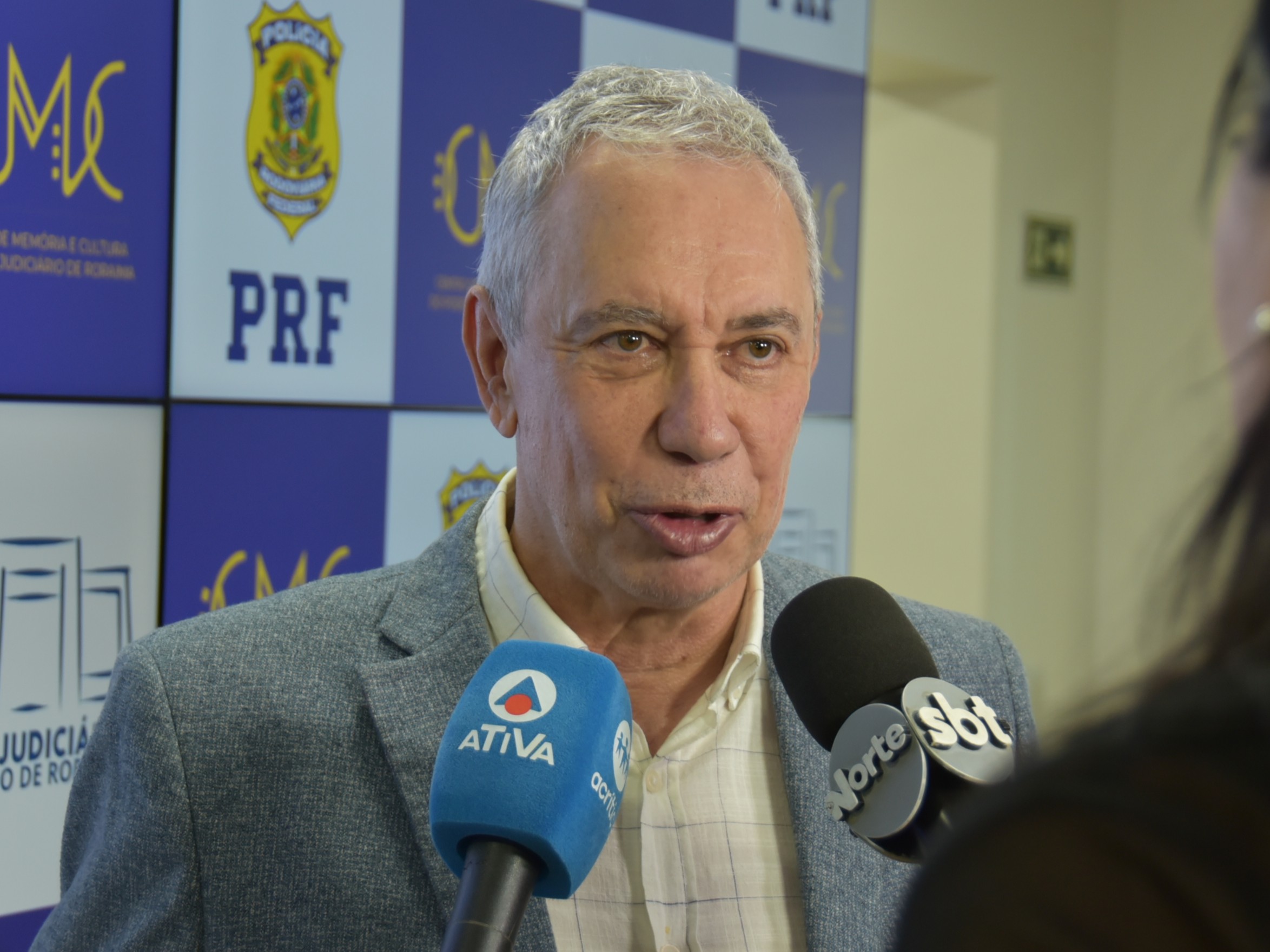 Imagem colorida mostra o vice-presidente do TJRR, desembargador Almiro Padilha, concedendo entrevista a emissoras de televisão, diante de painel com as identidades visuais da PRF e do Centro de Memória e Cultura.