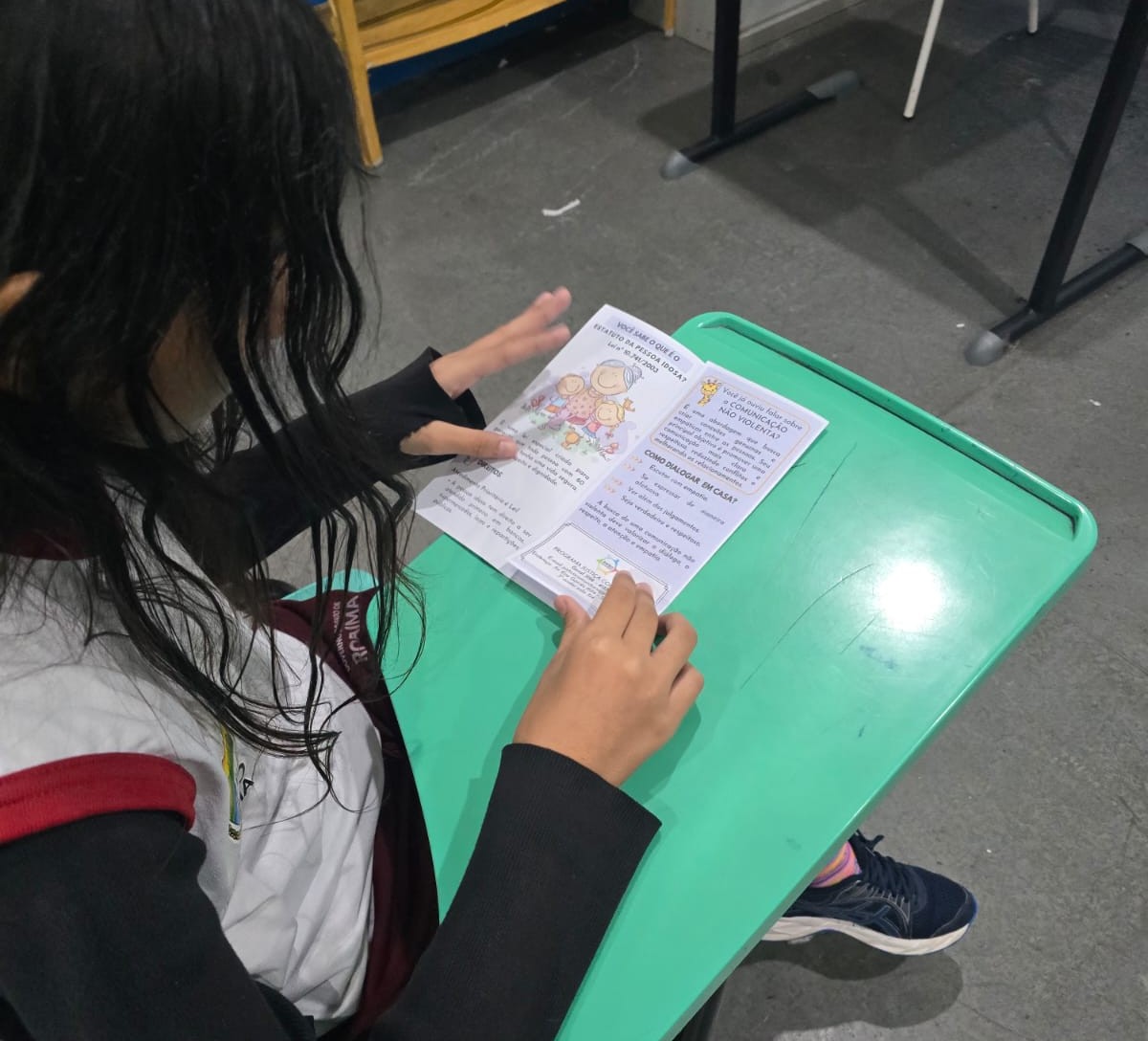 Uma estudante, sentada em uma carteira escolar verde, lê atentamente uma cartilha educativa sobre prevenção da violência. A imagem registra apenas parte do seu rosto e suas mãos segurando o material.