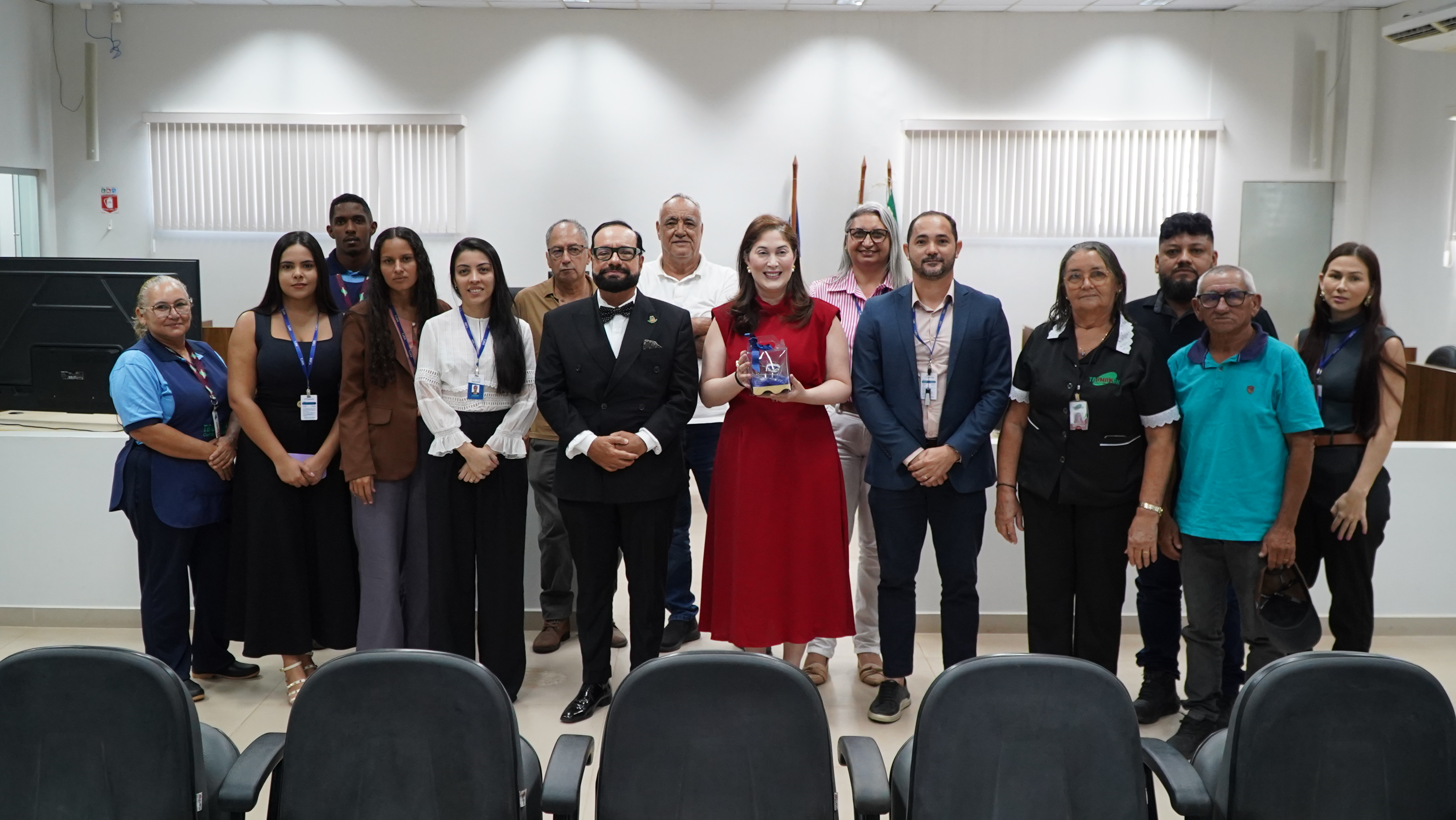 Imagem colorida registra servidores, magistrados e integrantes da comitiva posando para uma fotografia coletiva no auditório. Ao centro, uma mulher de vestido vermelho segura uma lembrança institucional.