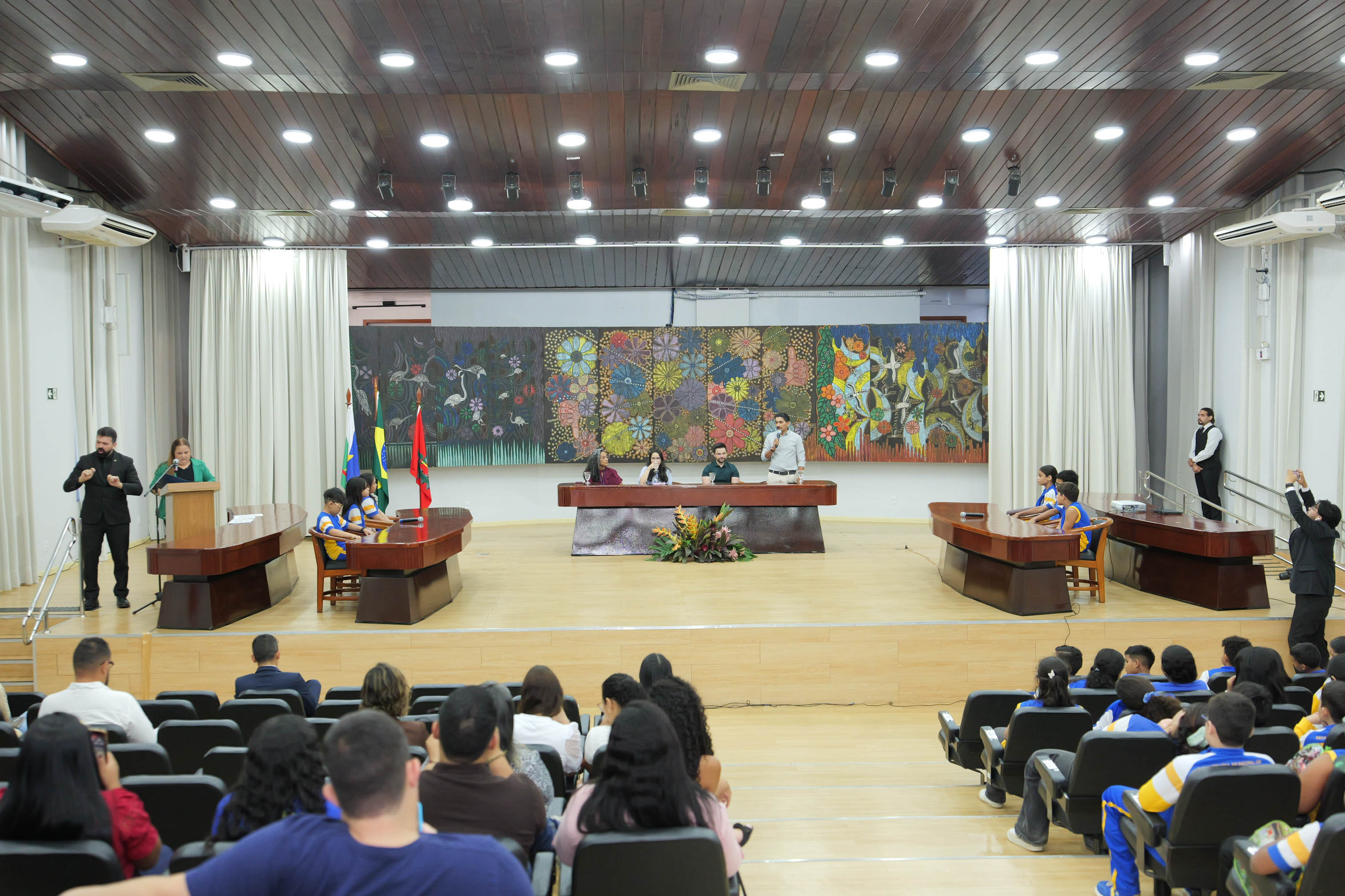 Visão ampla do auditório do Tribunal de Justiça de Roraima durante mesa compartilhada. No palco, estudantes participam, enquanto servidores conduzem a programação na mesa principal. A plateia é formada por alunos, professores e convidados. À esquerda, um intérprete de Libras realiza a tradução da atividade.