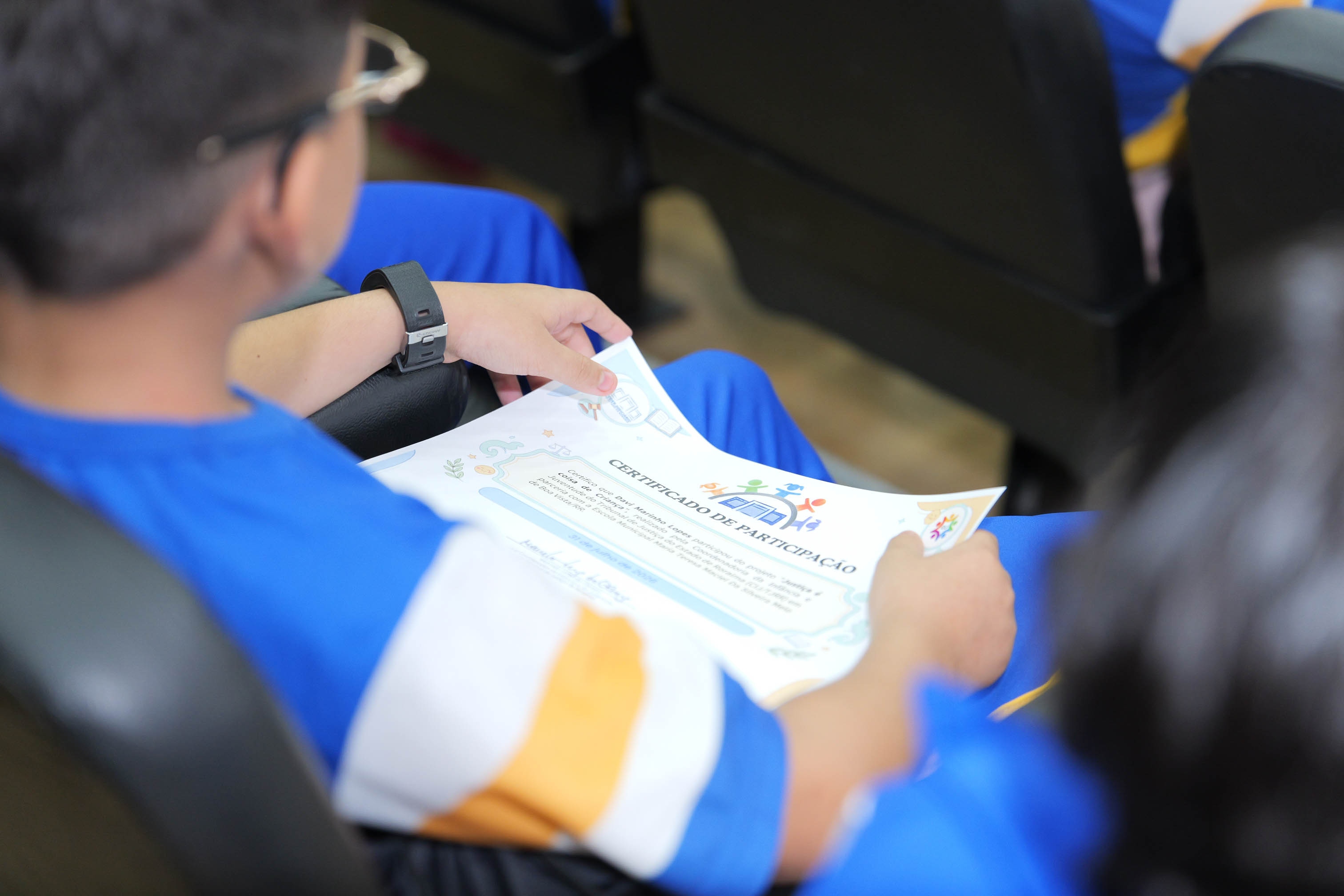  Em primeiro plano, um estudante sentado na plateia segura um certificado de participação com as duas mãos. Ele veste uniforme azul e usa relógio de pulso preto. Ao fundo, outros alunos aparecem desfocados acompanhando a programação.