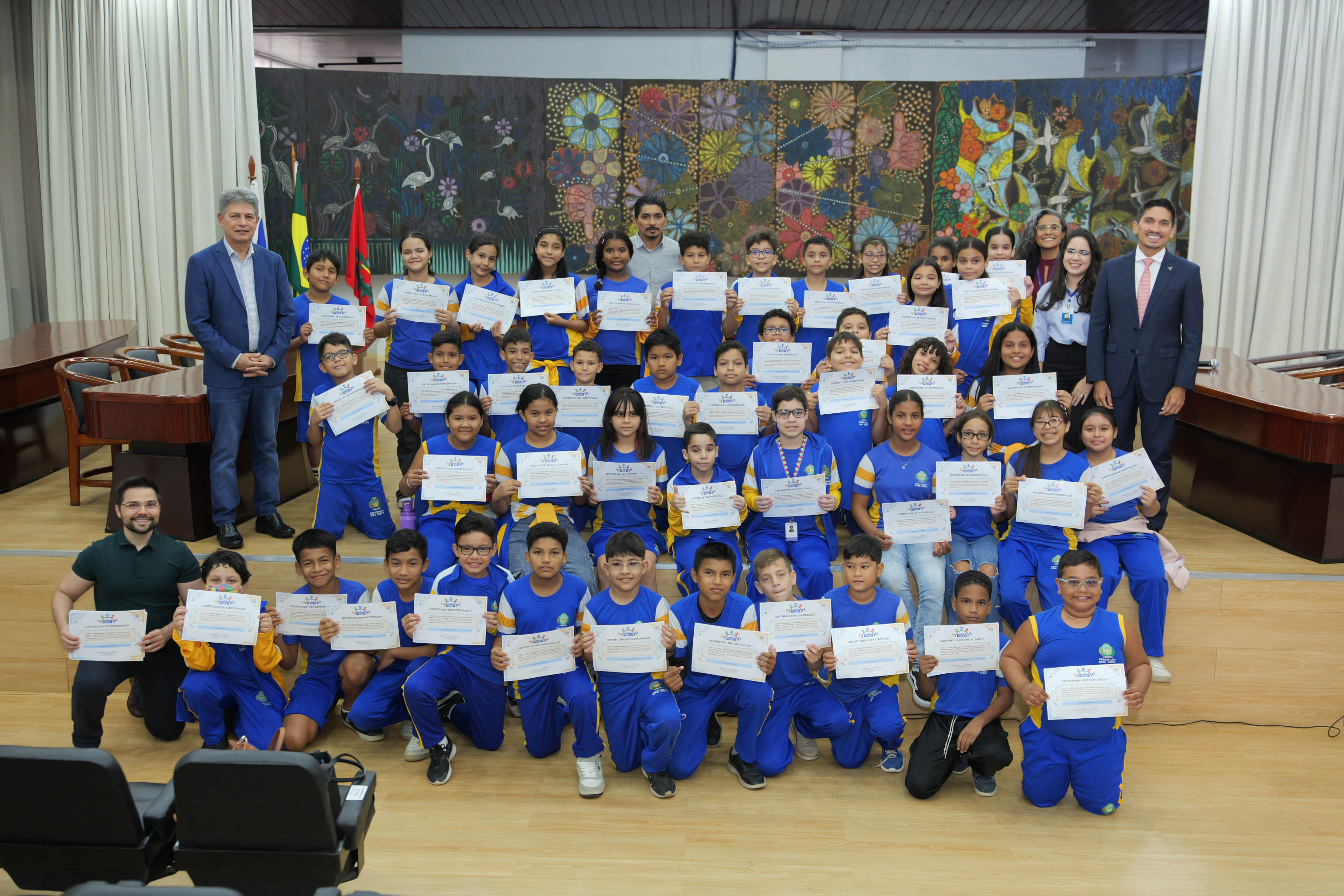 Foto oficial reúne dezenas de estudantes uniformizados segurando certificados de participação. Ao lado deles estão representantes do Tribunal de Justiça de Roraima, e o professor. O grupo posa em frente ao painel artístico do auditório.