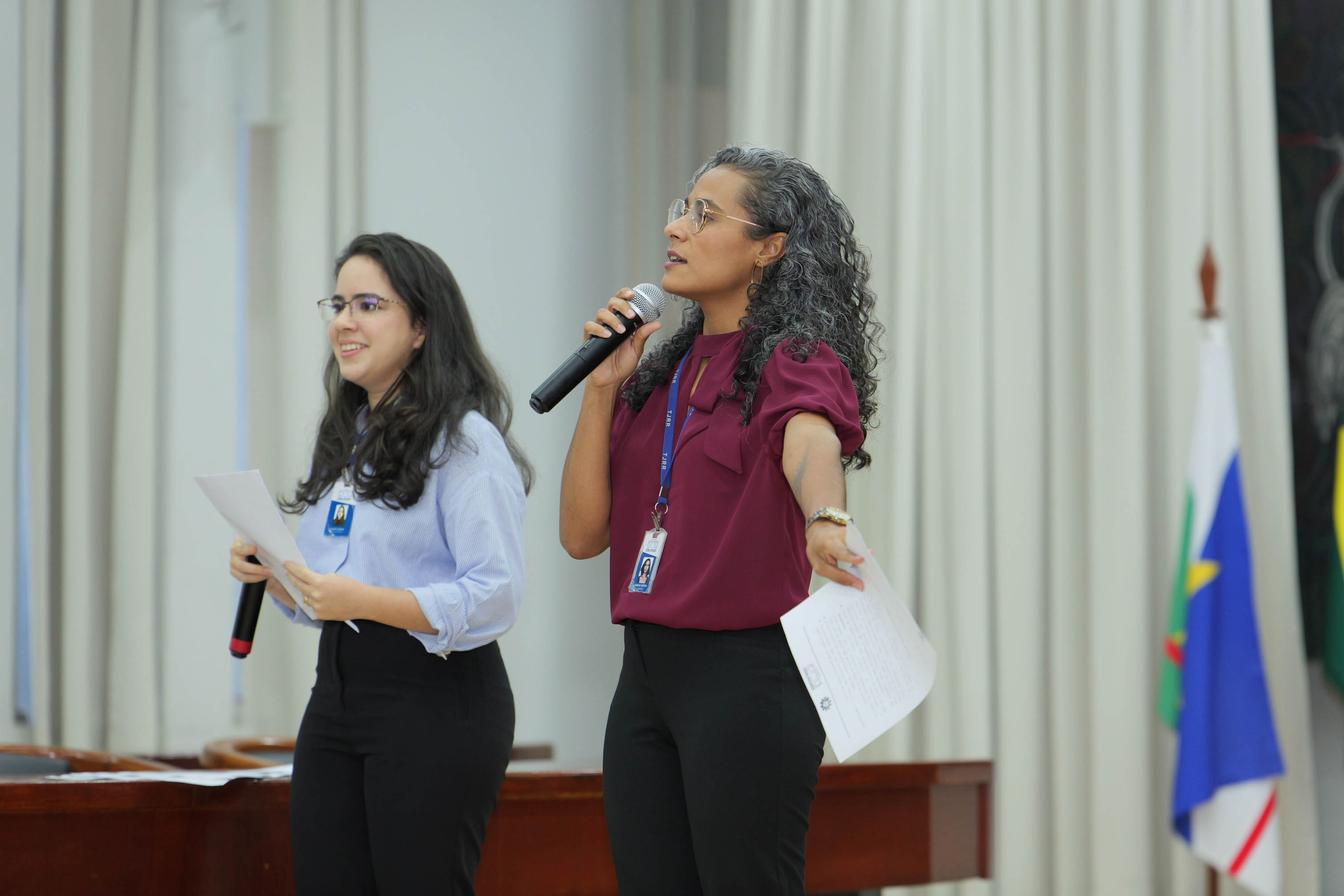 Duas servidoras conduzem relatório sobre as salas temáticas. Uma delas, de blusa vinho e crachá, fala ao microfone enquanto faz um gesto em direção ao público. Ao lado, outra servidora, de camisa azul-clara e óculos, segura um microfone e folhas com anotações.