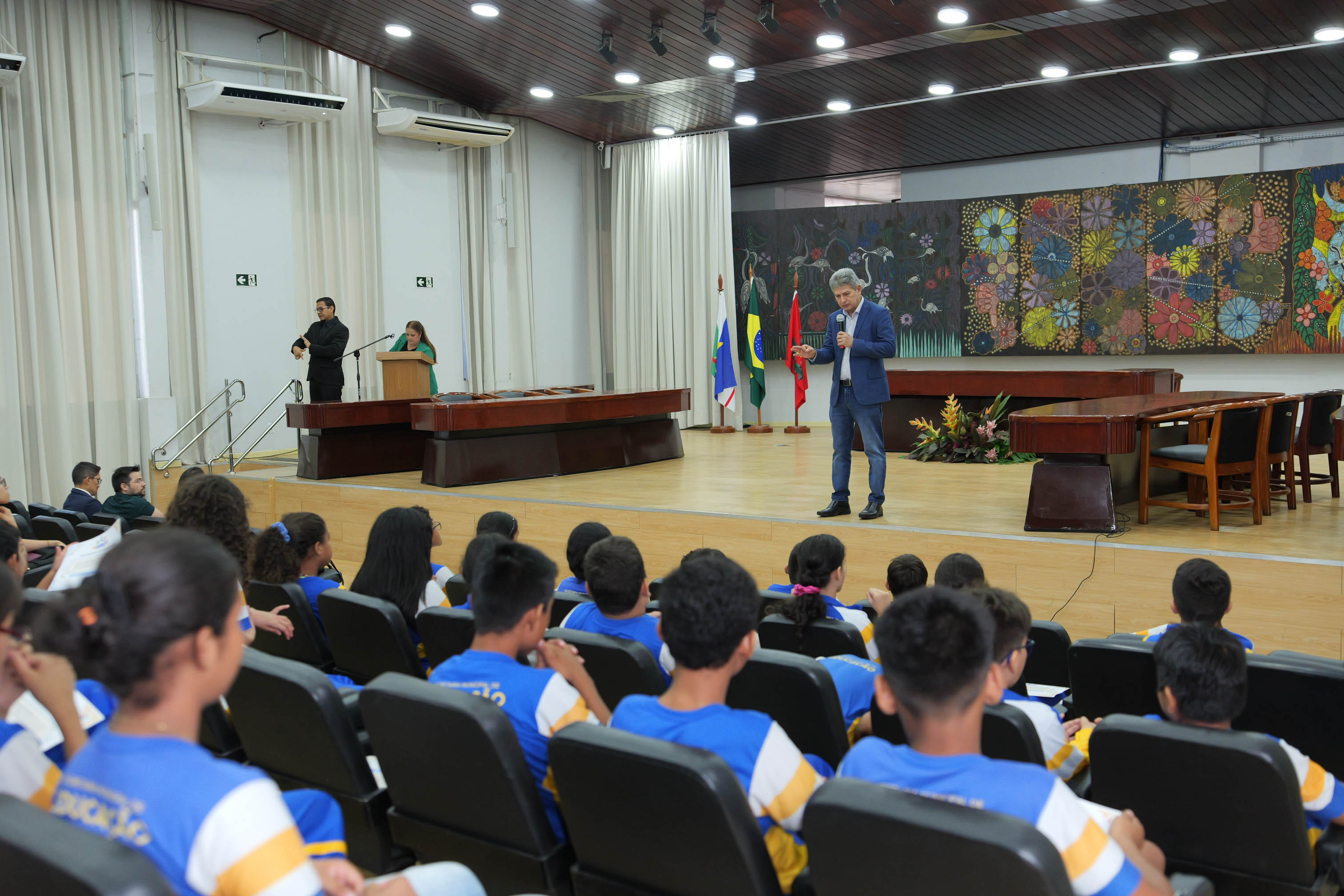 Juiz Parima Veras fala ao público no palco do auditório do Tribunal de Justiça de Roraima. À sua frente, estudantes uniformizados acompanham atentamente a apresentação. Ao lado do palco, um intérprete de Libras realiza a tradução da fala.