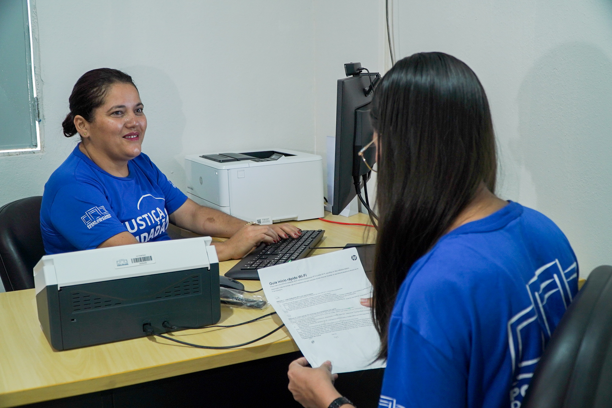 Fotografia colorida mostra duas servidoras vestindo camisetas azuis do programa Justiça Cidadã durante atendimento na nova unidade. Uma delas está sentada diante do computador, enquanto a outra, vista de costas, segura documentos. Sobre a mesa há teclado, monitor e impressoras.