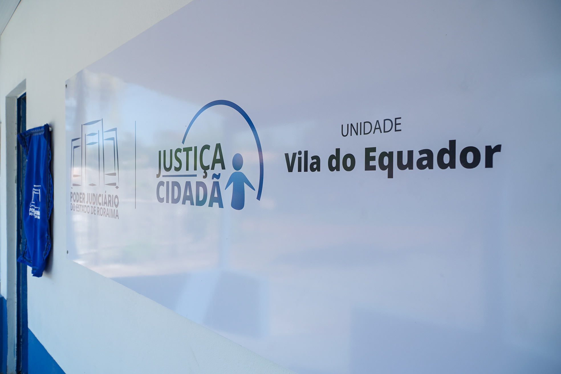 Fotografia colorida destaca a placa de identificação instalada na parede da unidade, com as marcas do Poder Judiciário do Estado de Roraima e do programa Justiça Cidadã, além do texto “Unidade Vila do Equador”.