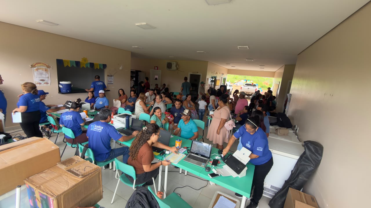 Interior movimentado com funcionários em camisetas azuis operando laptops e impressoras em mesas verdes. Grande público aguarda ao fundo e lateralmente. Caixas de suprimentos visíveis. Interior movimentado com funcionários em camisetas azuis operando laptops e impressoras em mesas verdes. Grande público aguarda ao fundo e lateralmente. Caixas de suprimentos visíveis.