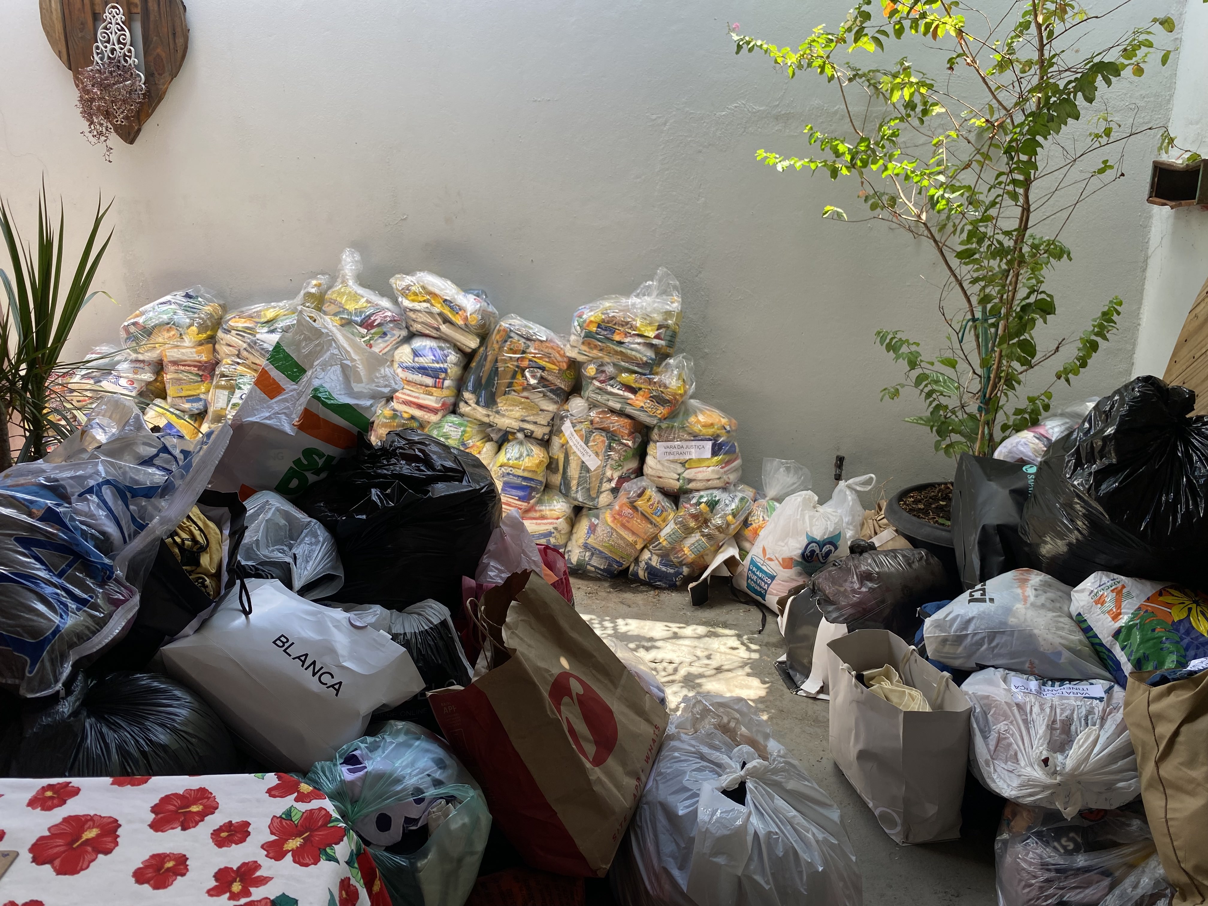 Fotografia de uma grande quantidade de doações arrecadadas, com cestas básicas de alimentos e sacos com suprimentos empilhados ao lado de uma parede externa. Fotografia de uma grande quantidade de doações arrecadadas, com cestas básicas de alimentos e sacos com suprimentos empilhados ao lado de uma parede externa.