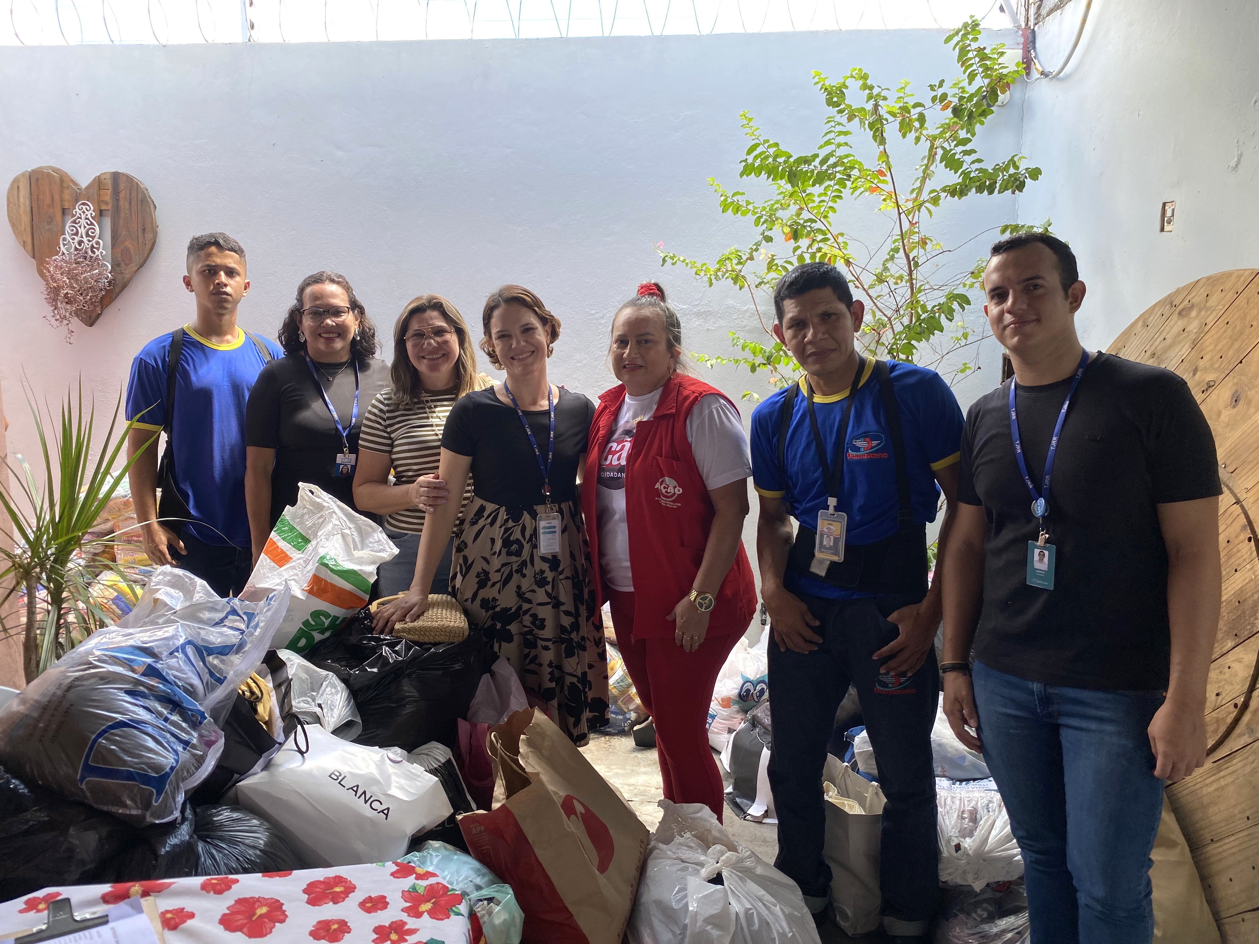 Fotografia de grupo de voluntários e servidores posando ao lado de uma grande pilha de doações, incluindo caixas, sacolas e pacotes. Fotografia de grupo de voluntários e servidores posando ao lado de uma grande pilha de doações, incluindo caixas, sacolas e pacotes.