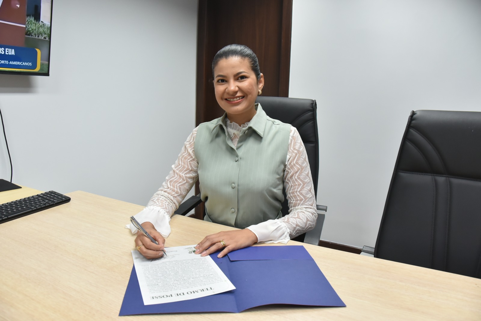 Fotografia colorida mostra uma mulher sorrindo enquanto assina o "Termo de Posse" sobre uma mesa de madeira clara. Ela usa blusa branca de mangas rendadas com colete bege-esverdeado e tem os cabelos presos. À sua frente está a pasta azul do TJRR aberta. Fotografia colorida mostra uma mulher sorrindo enquanto assina o "Termo de Posse" sobre uma mesa de madeira clara. Ela usa blusa branca de mangas rendadas com colete bege-esverdeado e tem os cabelos presos. À sua frente está a pasta azul do TJRR aberta.