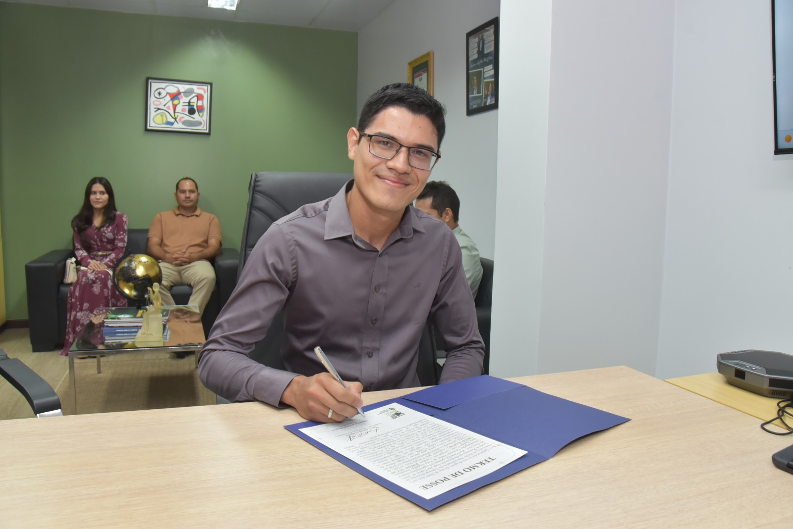 Fotografia colorida mostra um homem de óculos e camisa cinza de manga longa sorrindo para a câmera enquanto assina o "Termo de Posse" sobre uma mesa de madeira clara. Ao fundo, em um sofá, observam uma mulher de vestido estampado e um homem de camisa polo clara. Fotografia colorida mostra um homem de óculos e camisa cinza de manga longa sorrindo para a câmera enquanto assina o "Termo de Posse" sobre uma mesa de madeira clara. Ao fundo, em um sofá, observam uma mulher de vestido estampado e um homem de camisa polo clara.