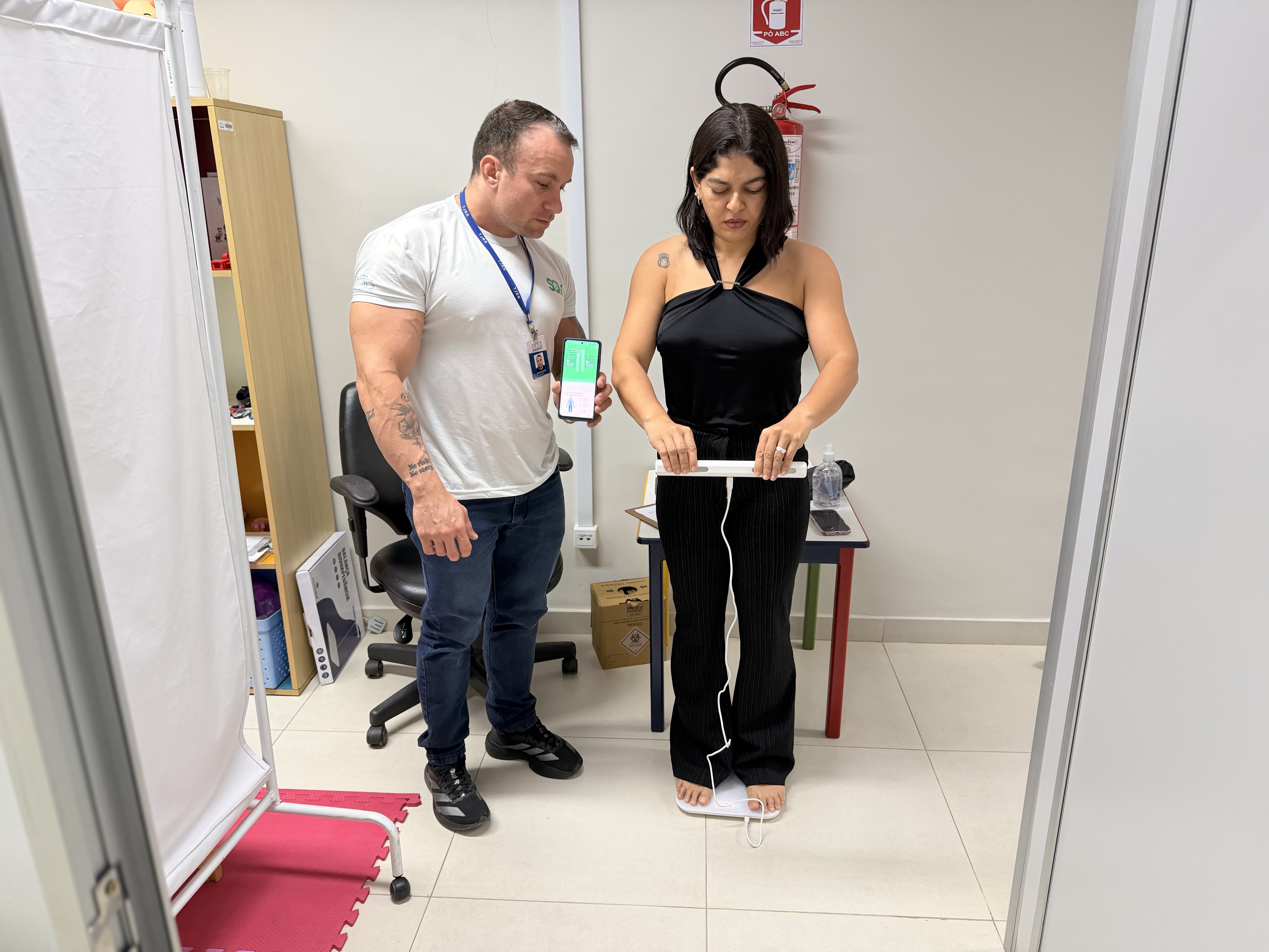 Uma mulher realiza exame de bioimpedância sobre uma balança digital, segurando os eletrodos com as mãos. Ao seu lado, um profissional segura um aparelho que exibe os resultados da medição em tempo real.